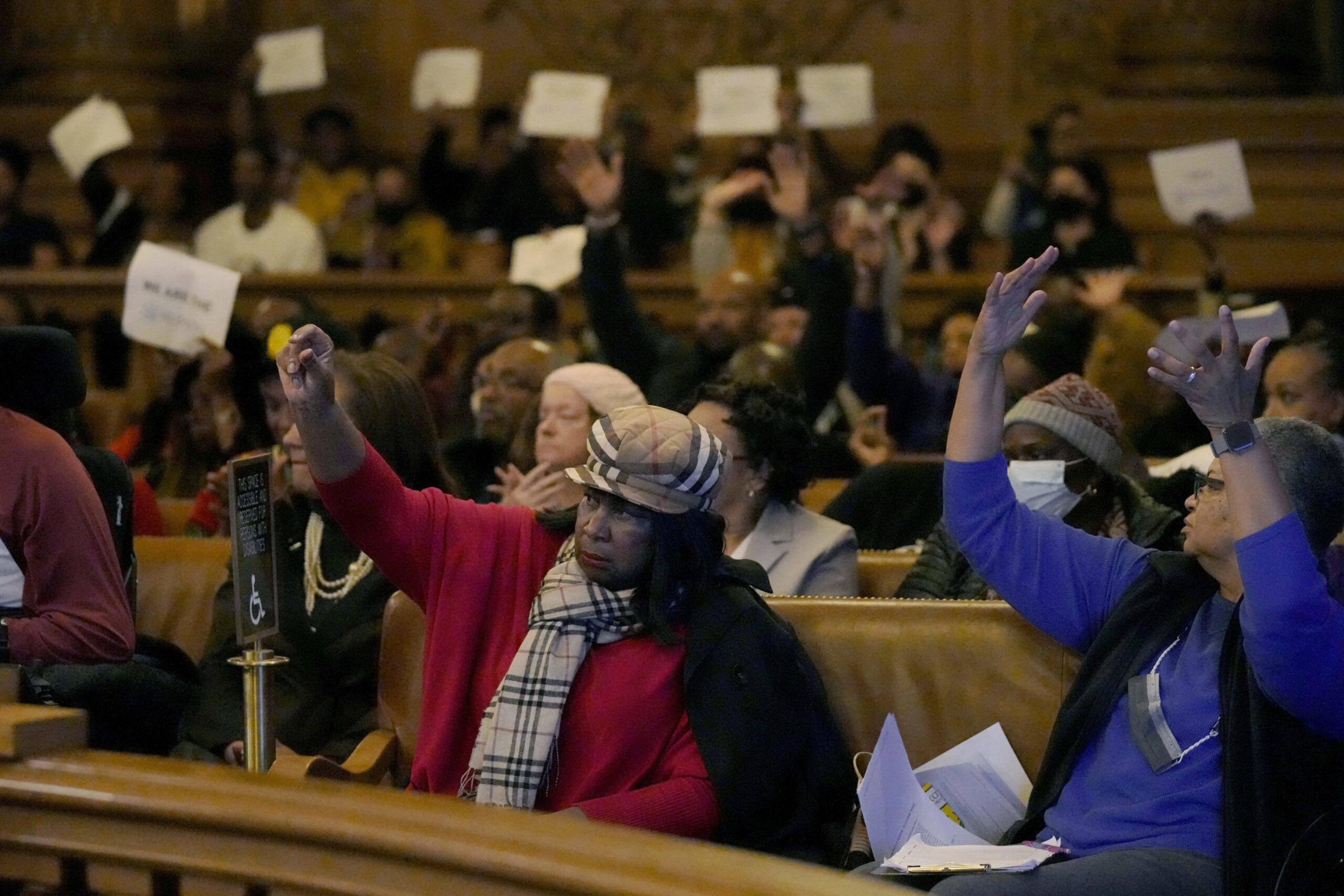 A diverse group of people sit in an auditorium, many raising their hands, with some holding up white signs.
