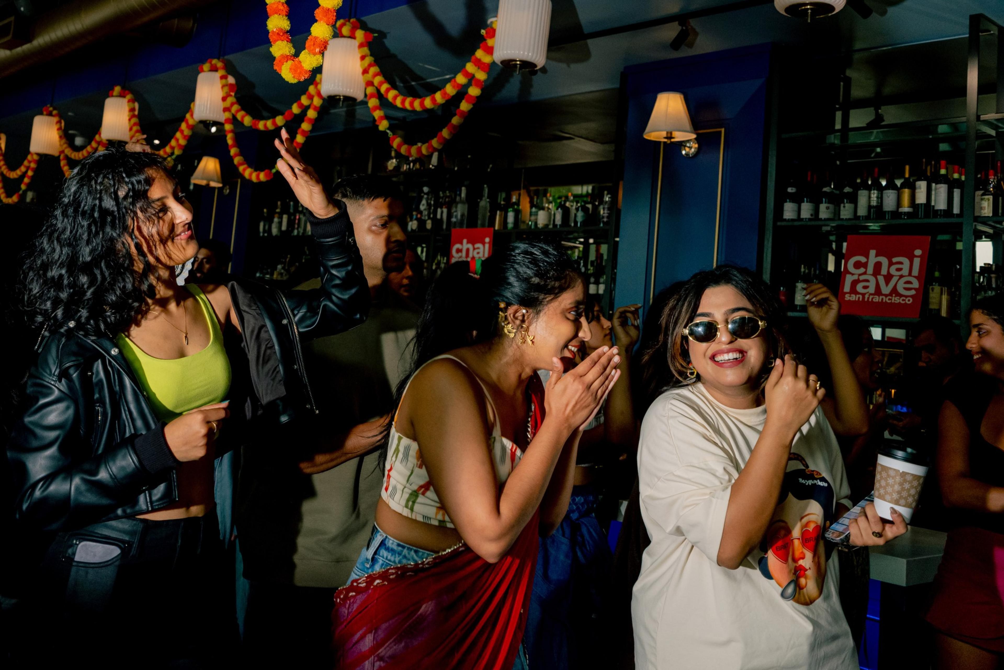A group of people enjoy a lively party with colorful decorations; one woman laughs clapping hands, another wears sunglasses and holds a coffee cup.
