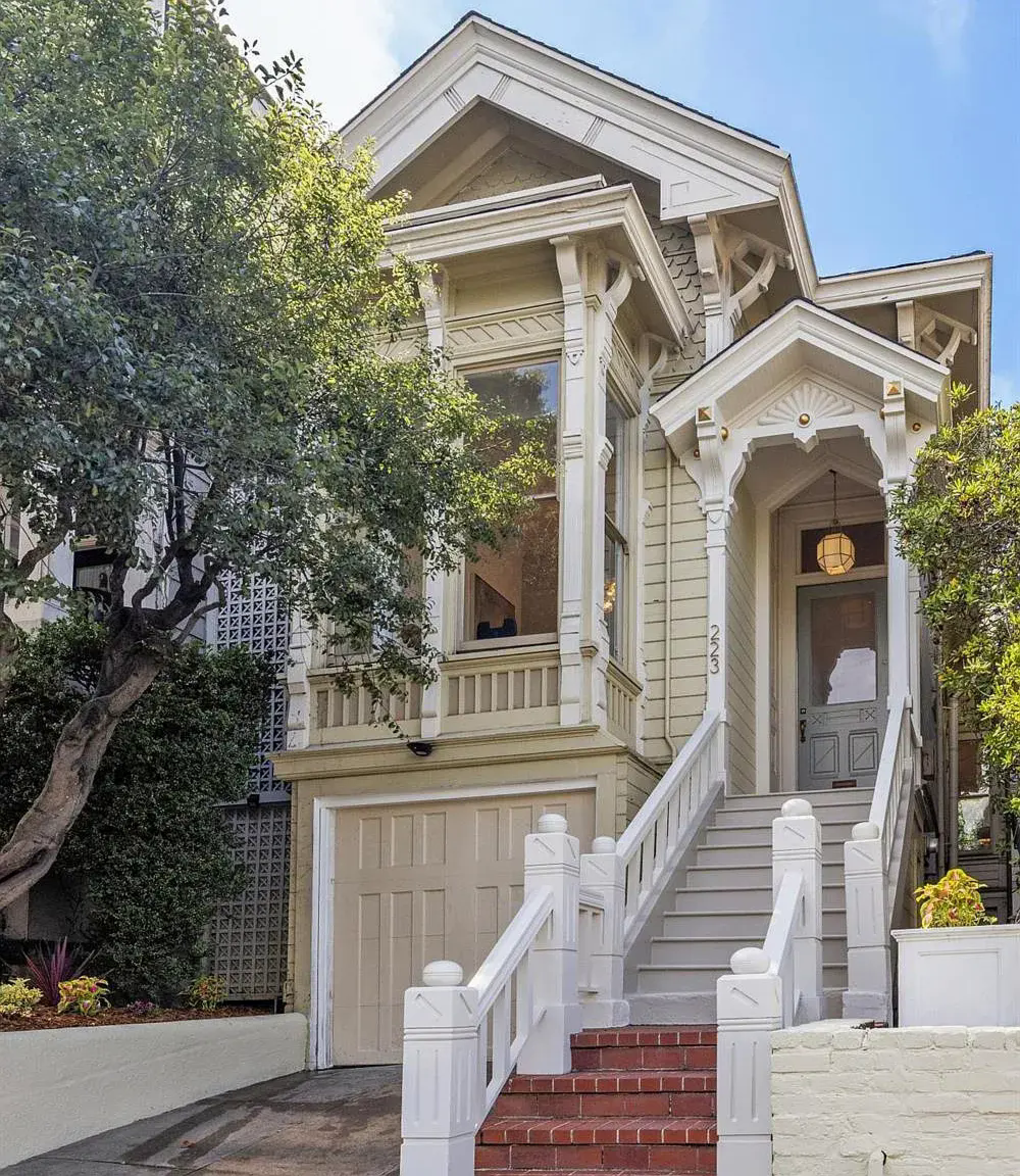 A light beige Victorian-style house with white trim, a front staircase, a garage, and a large tree partially shading the left side.
