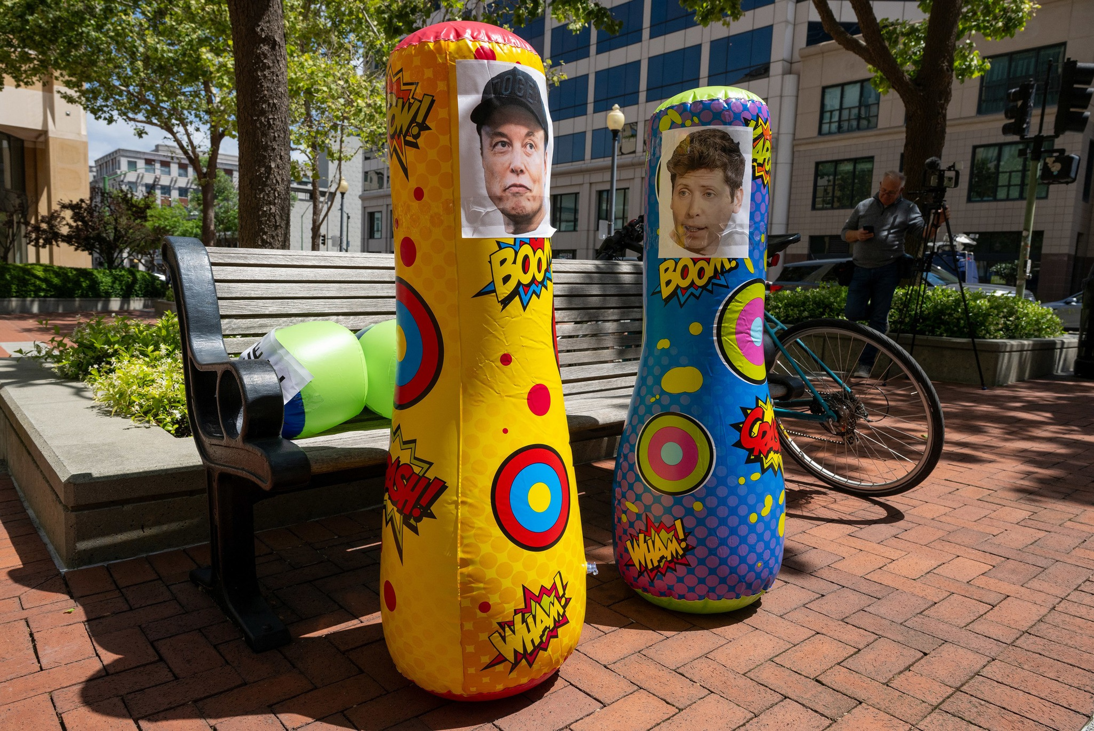 Two colorful inflatable punching bags with photos of a man and a woman taped on top stand on a brick sidewalk near a bench and a bicycle.