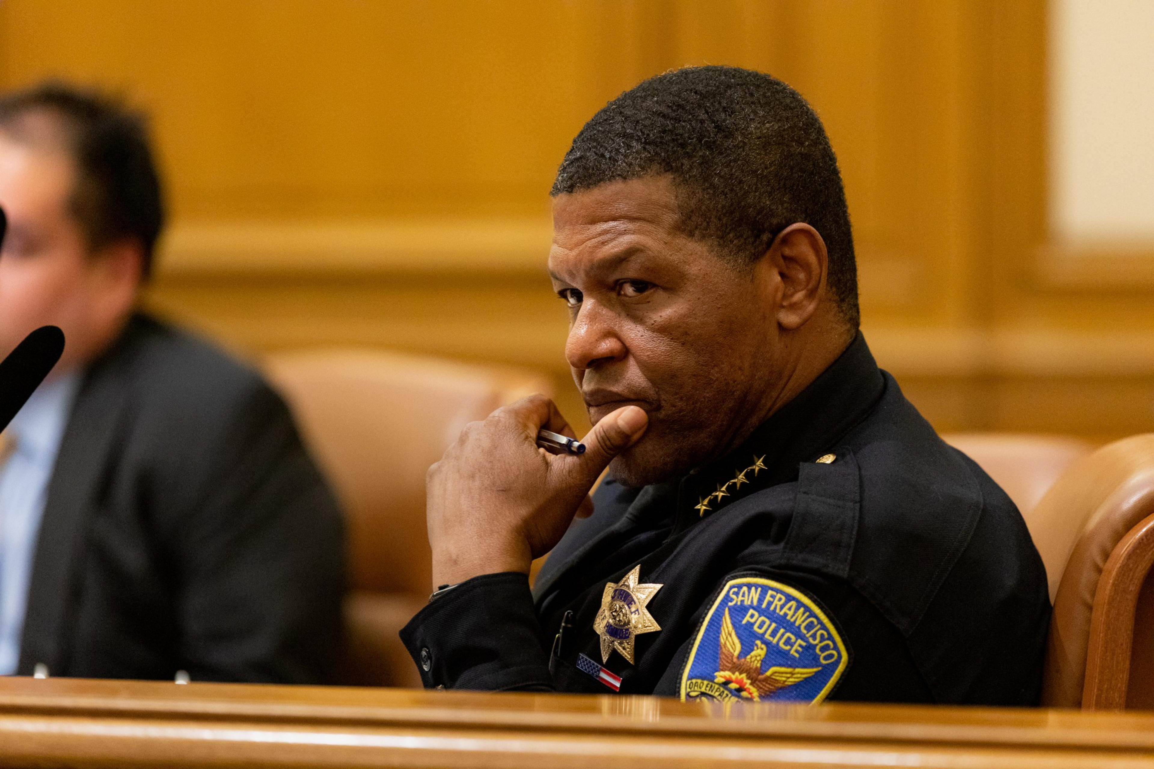 A man in a San Francisco Police uniform sits thoughtfully with his hand near his mouth during a meeting or hearing.