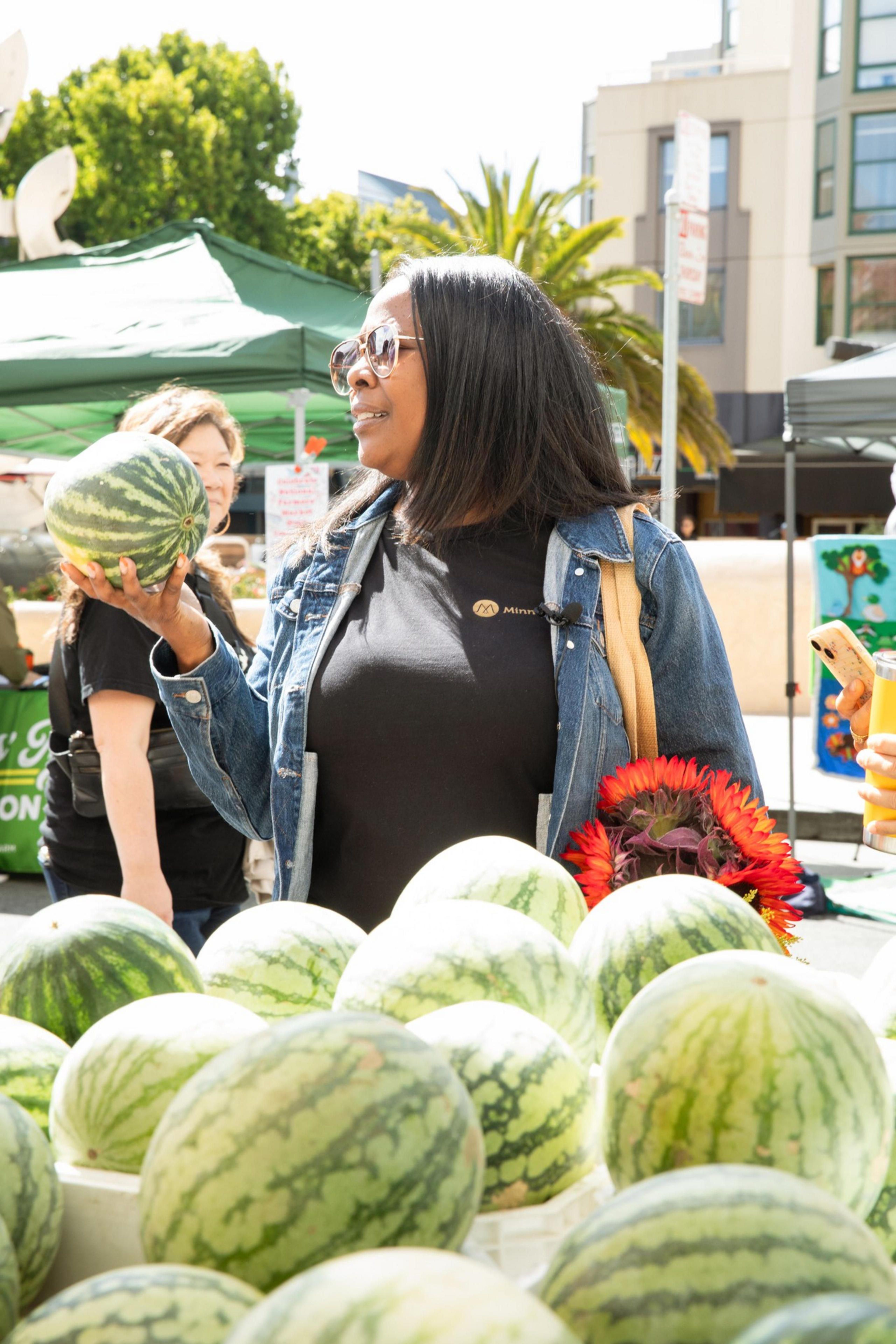 A woman is holding a watermelon at an outdoor market. She wears sunglasses and a denim jacket, with another watermelon in front of her and sunflowers in her tote bag.