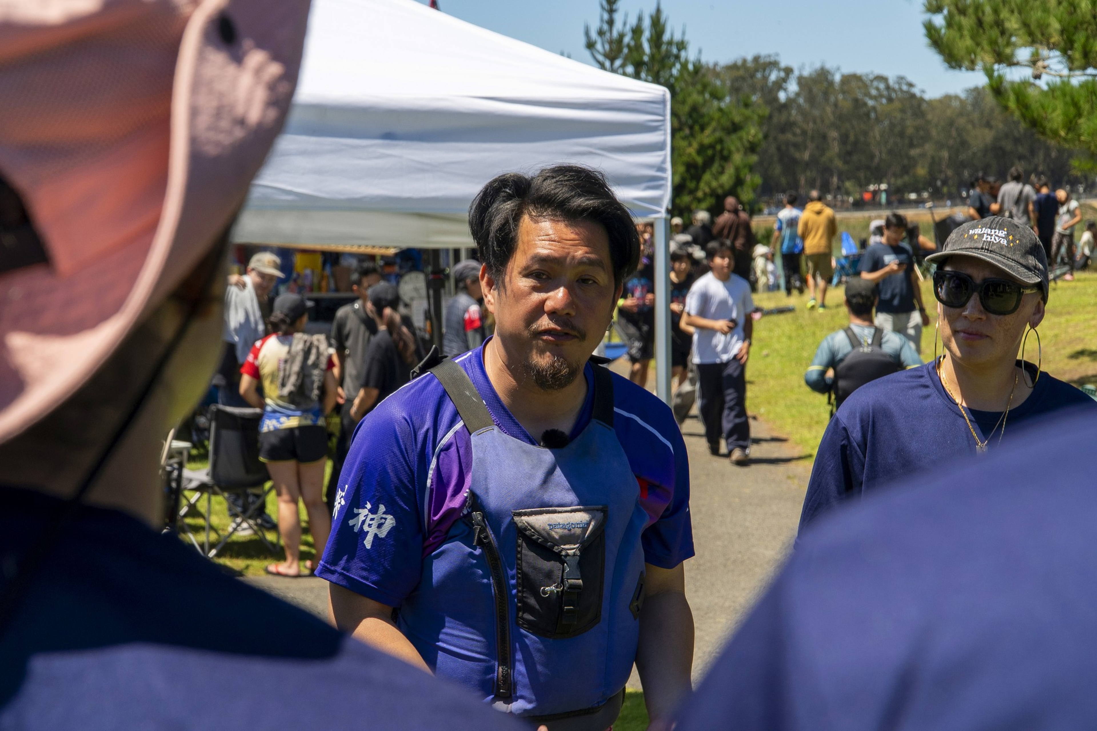 A man wearing a blue shirt and chest pack stands outside talking, with people and tents in the sunny park background.