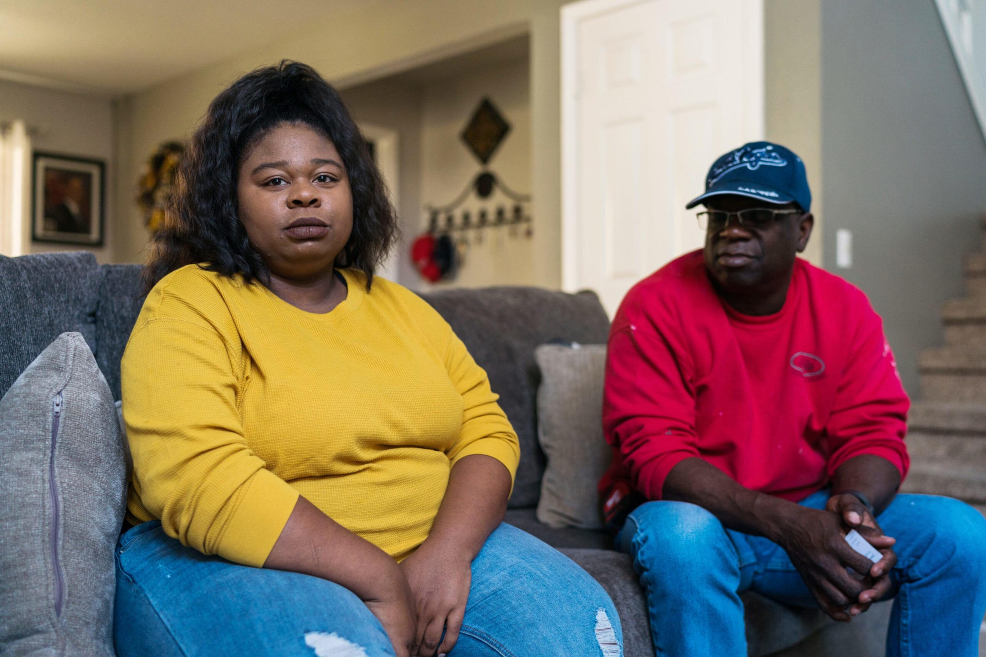 A woman in a yellow sweater and ripped jeans sits on a gray couch next to a man in a red sweatshirt, blue cap, and glasses looking at her.