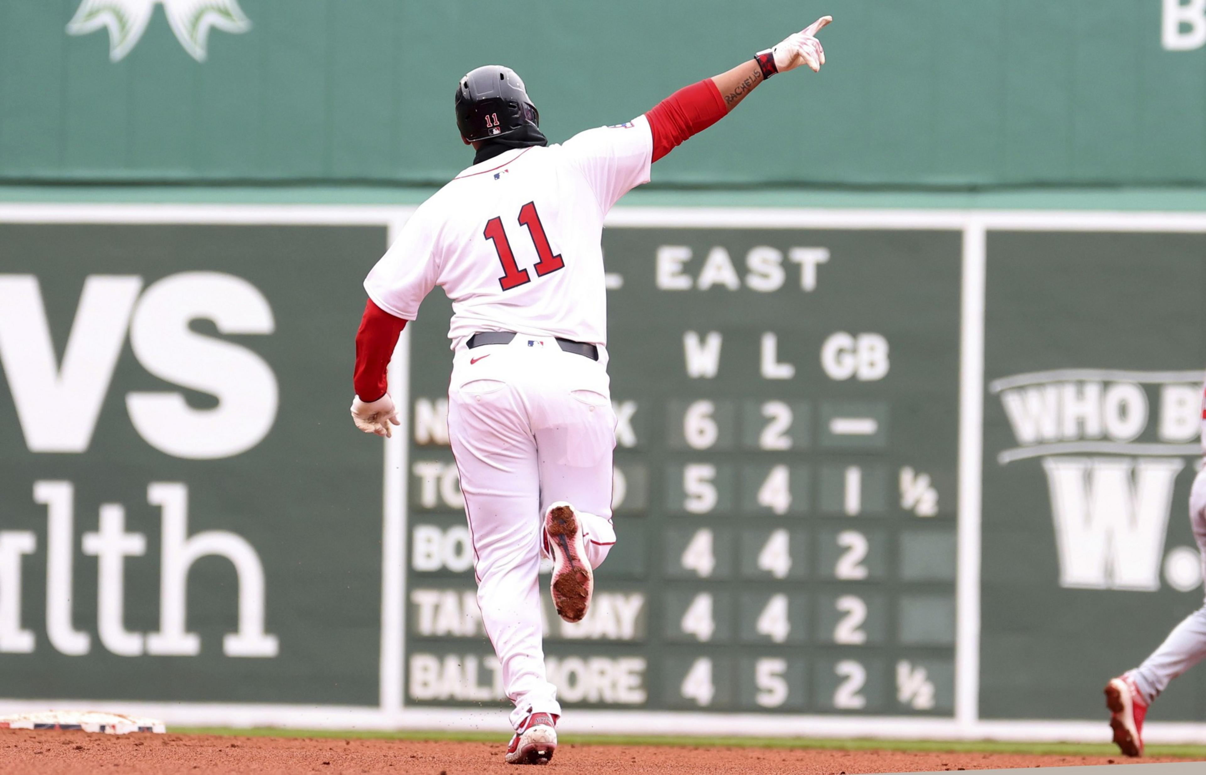 A baseball player in a white uniform with number 11 runs on a field, pointing upwards. Behind is a green scoreboard displaying team standings.