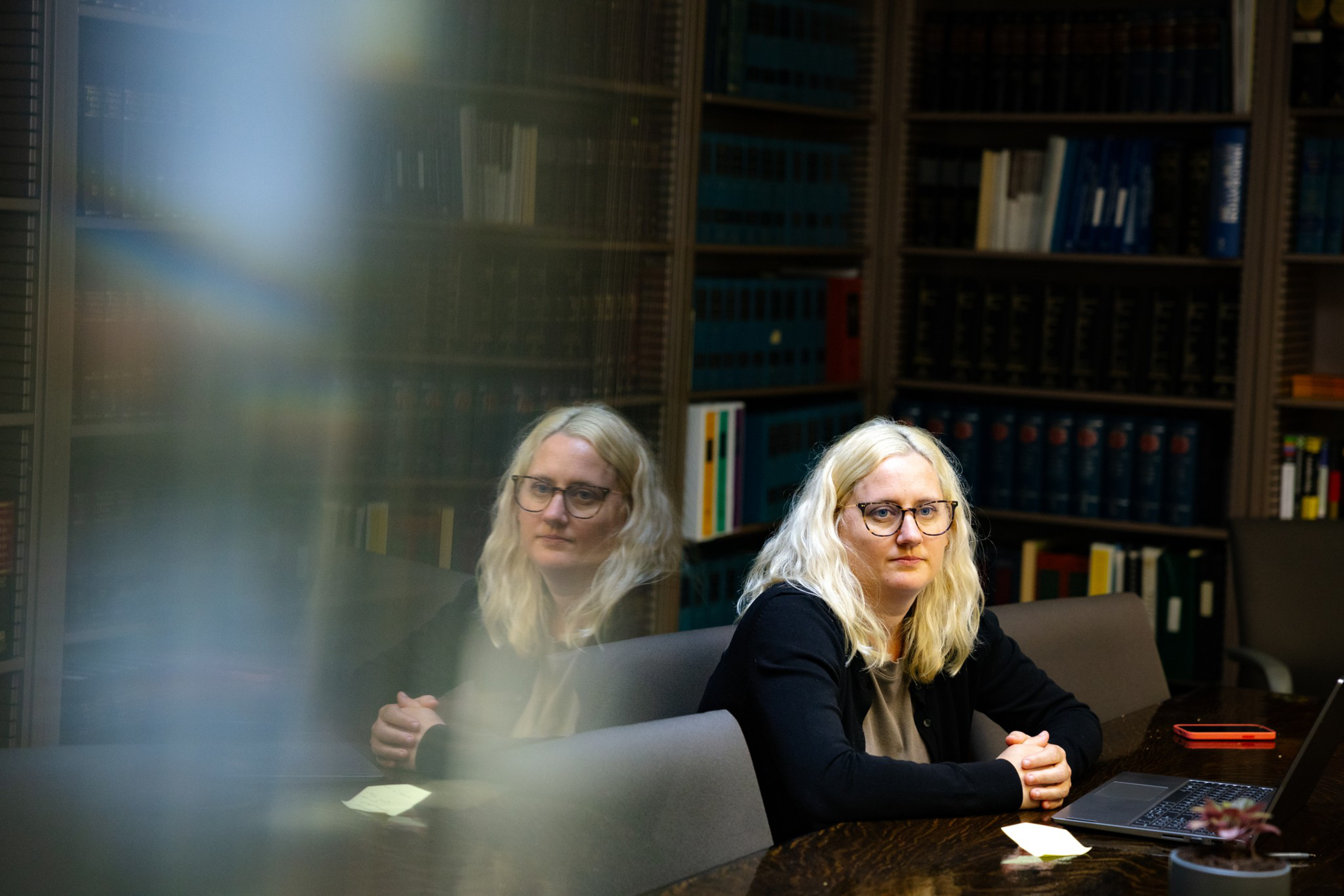 A woman with blonde hair and glasses sits at a conference table with a laptop, folded hands, and a reflective glass wall showing her mirrored image.