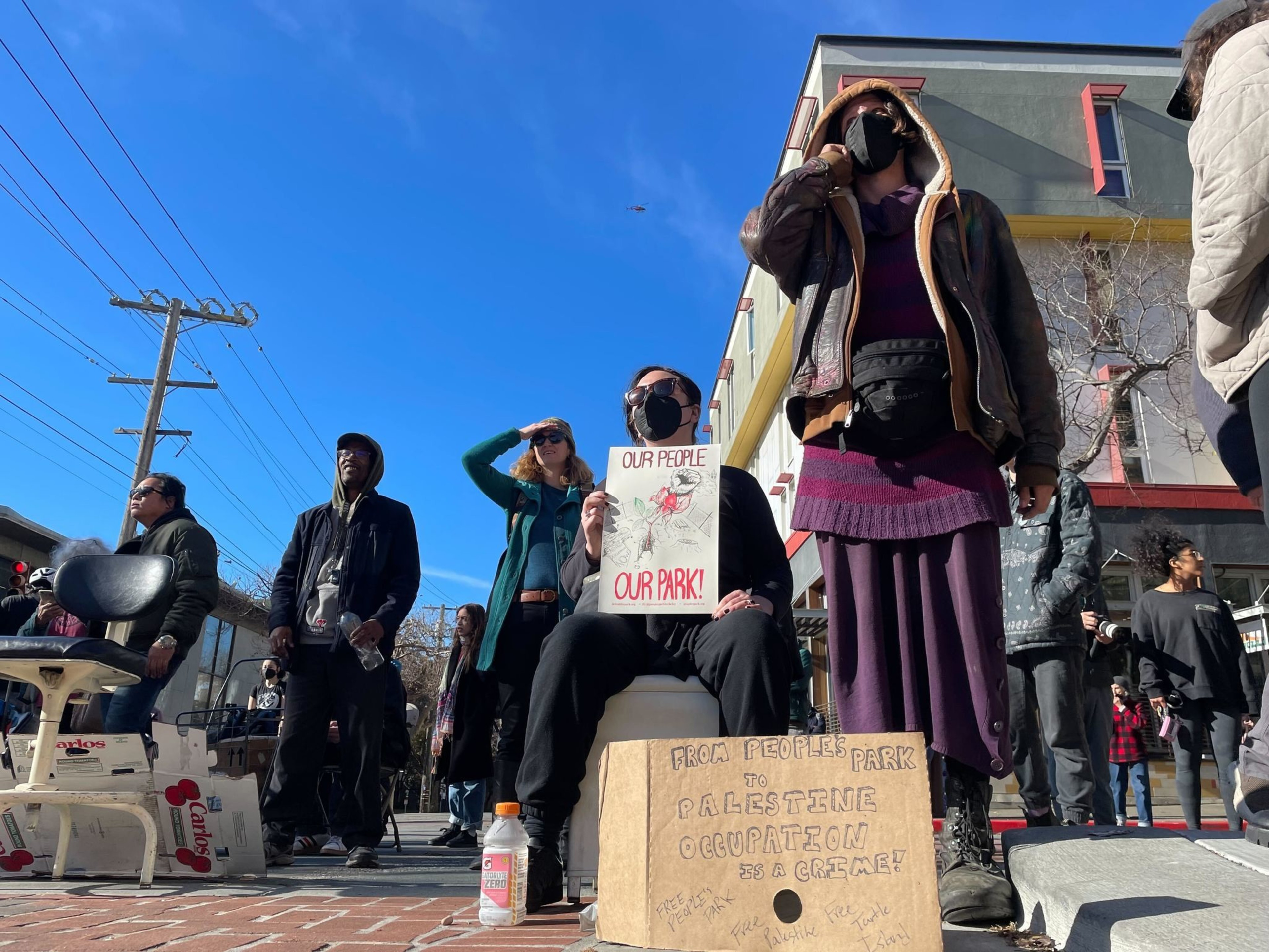Several mask-wearing people in dark colored clothing stand and hold signs at a city intersection under sunny blue sky.
