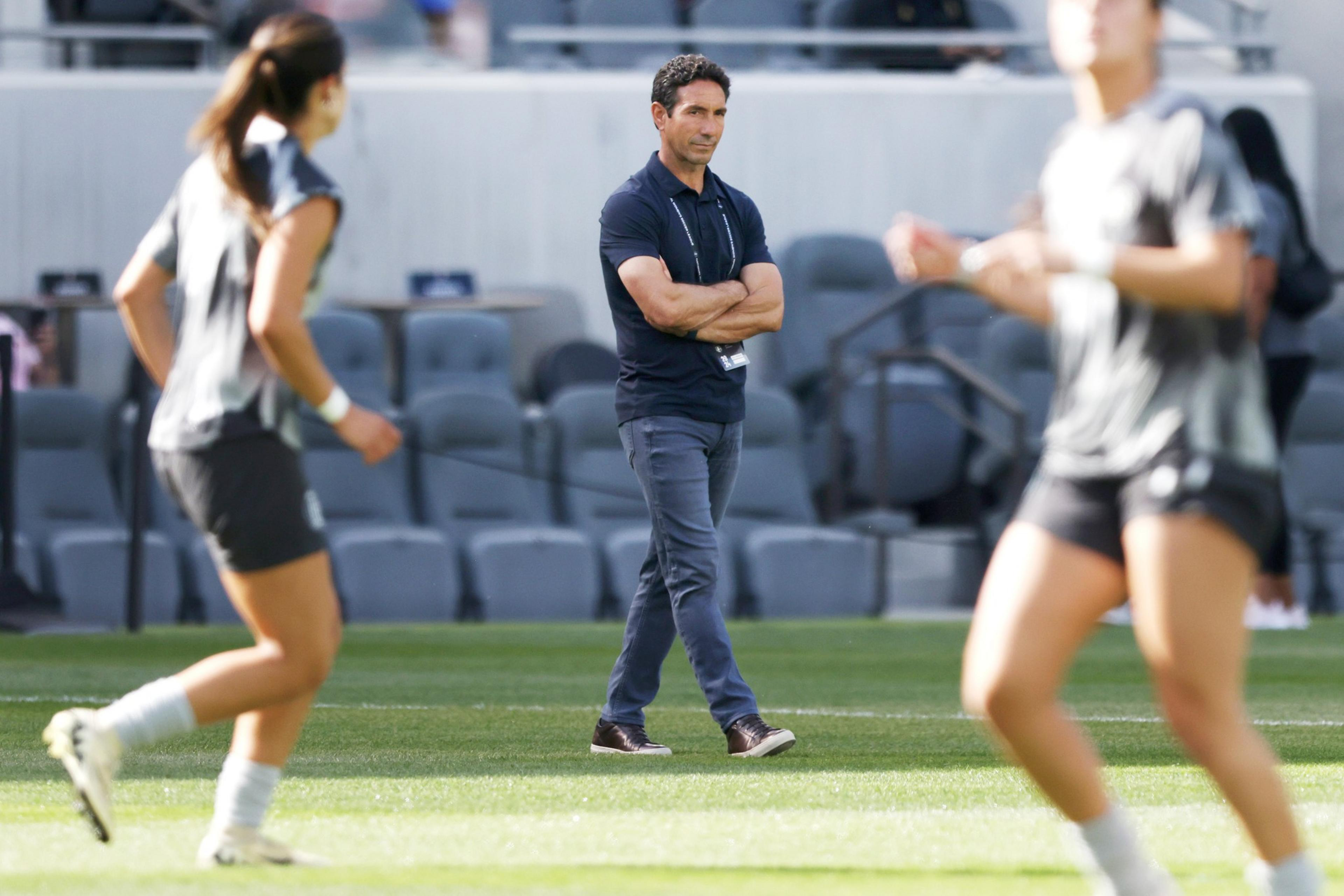 A man stands with arms crossed watching blurred figures of athletes training on a field.