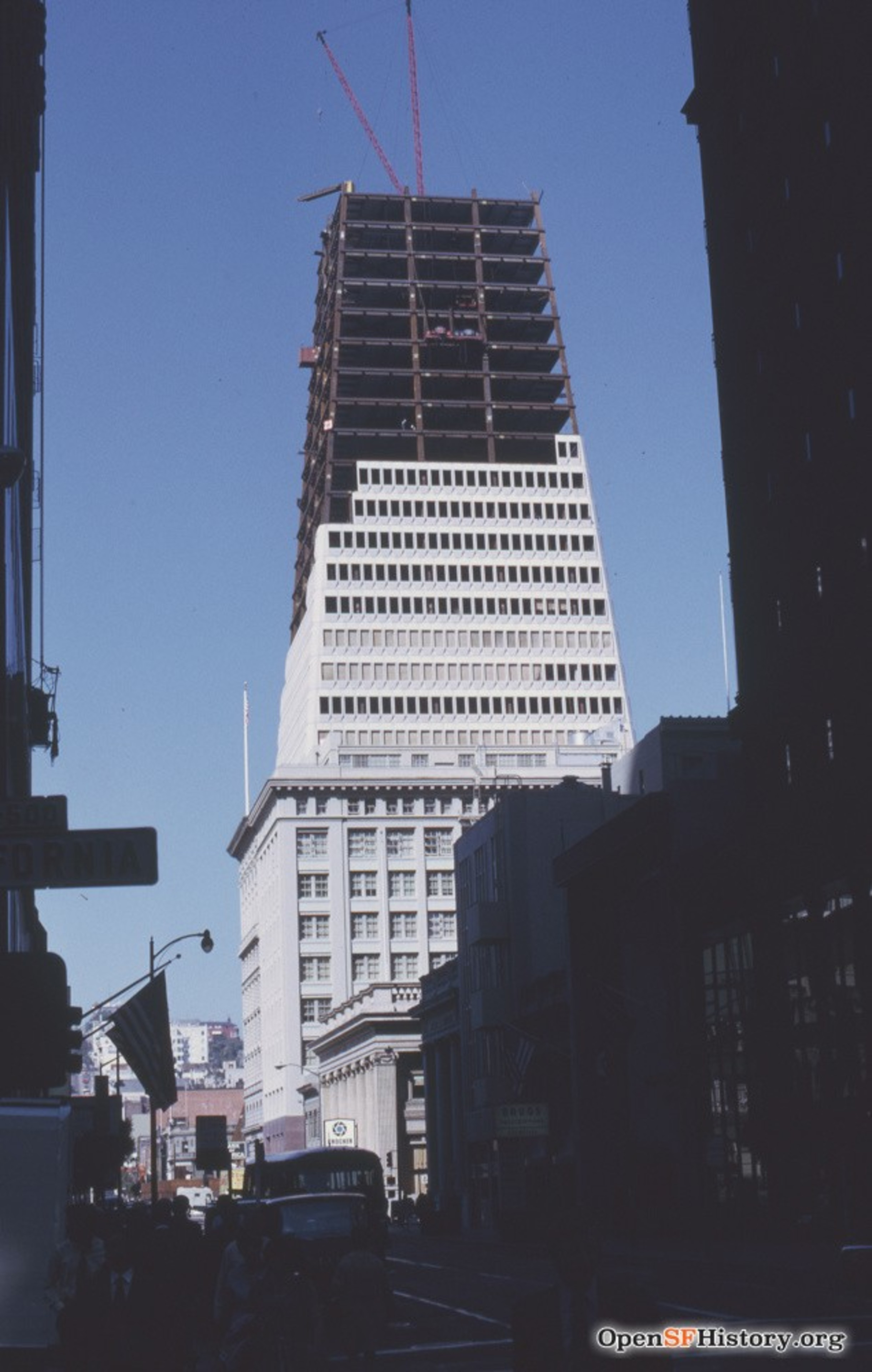 A skyscraper under construction looms over a bustling city street with pedestrians and vintage cars.