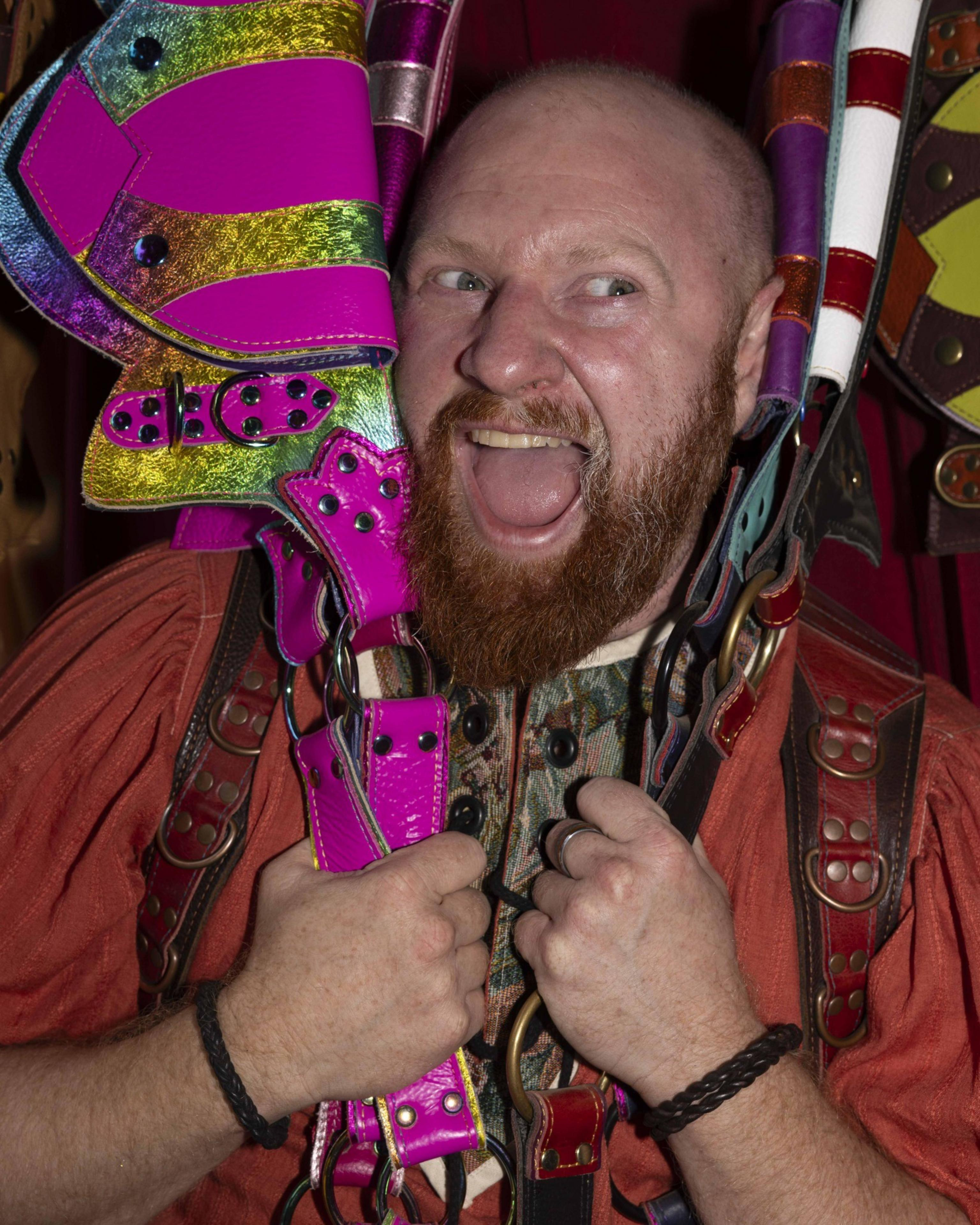 A man with a red beard grins widely while holding colorful, shiny leather straps, including pink and rainbow ones, against his head and shoulders.