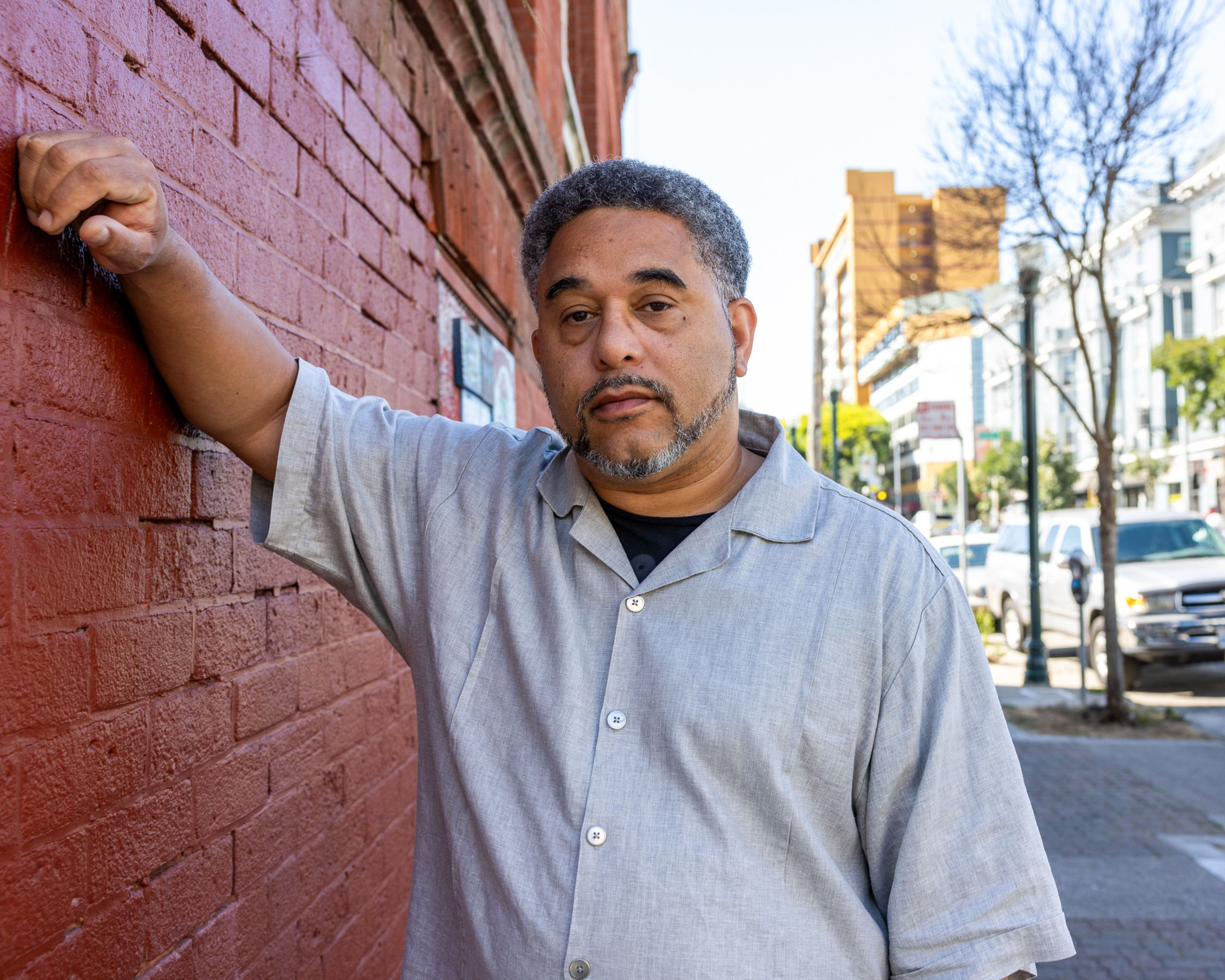 A man in a light gray shirt leans against a red brick wall on a city street, with buildings, parked cars, and leafless trees in the background.