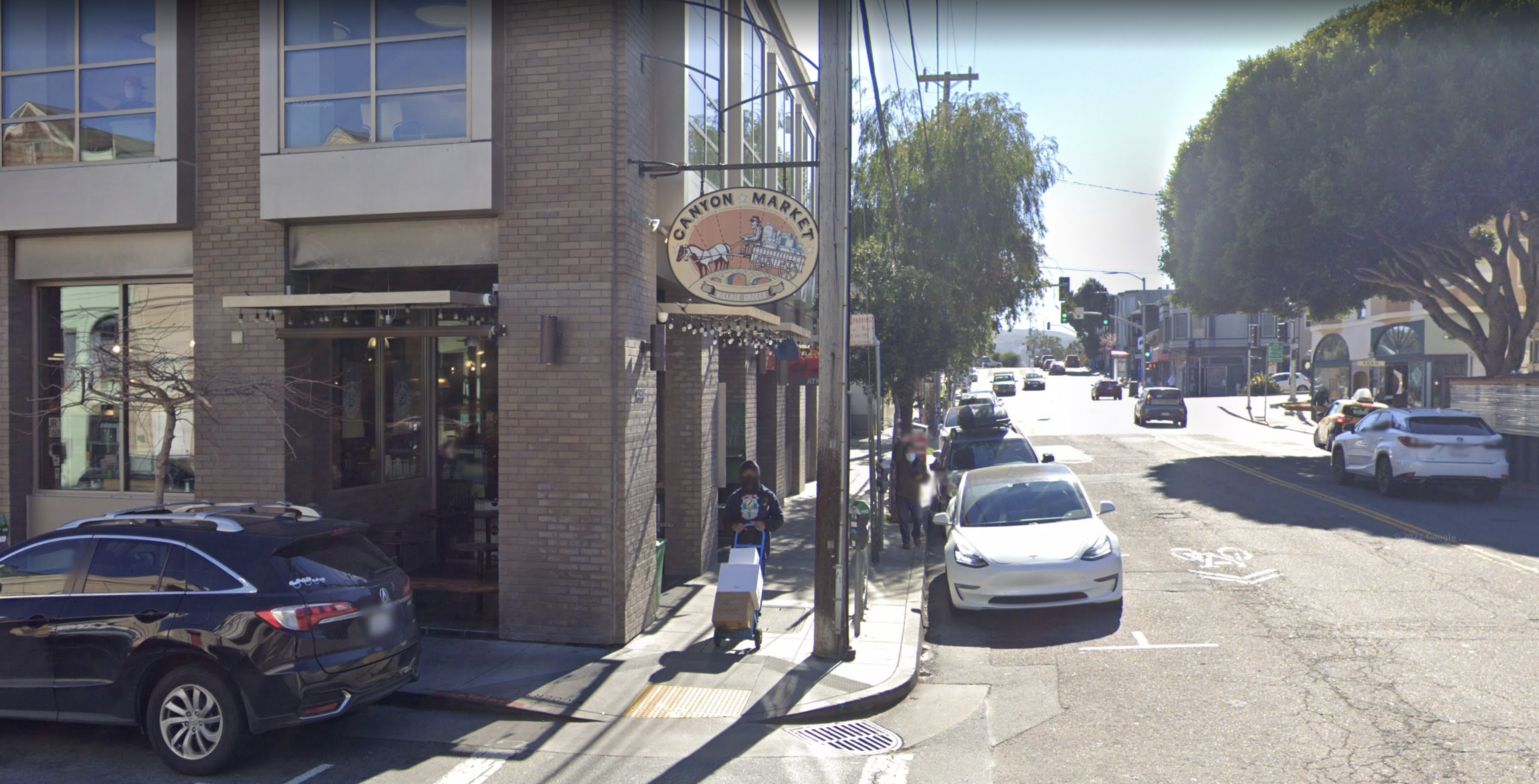 A brick building with a "Canyon Market" sign stands next to a sidewalk where a person pushes a dolly; cars are parked along a sunlit street lined with trees.