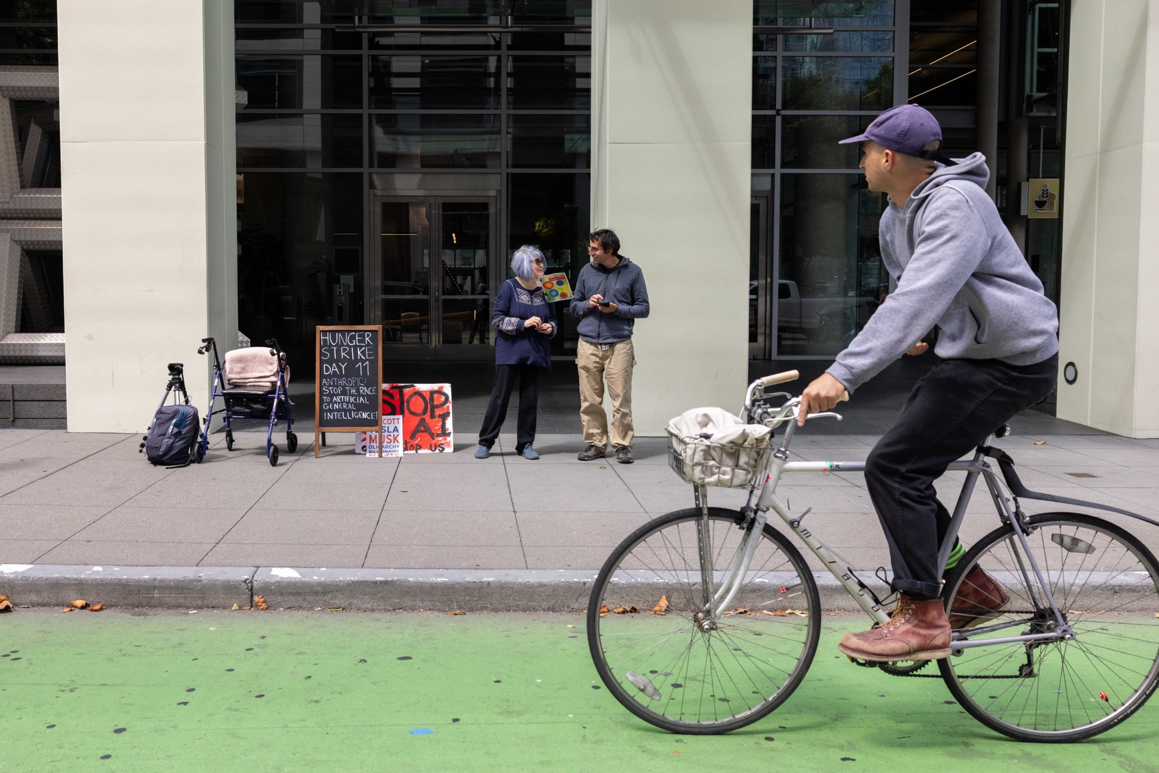 A person wearing a helmet rides a bicycle on a path in front of a building with large windows and some outdoor seating.