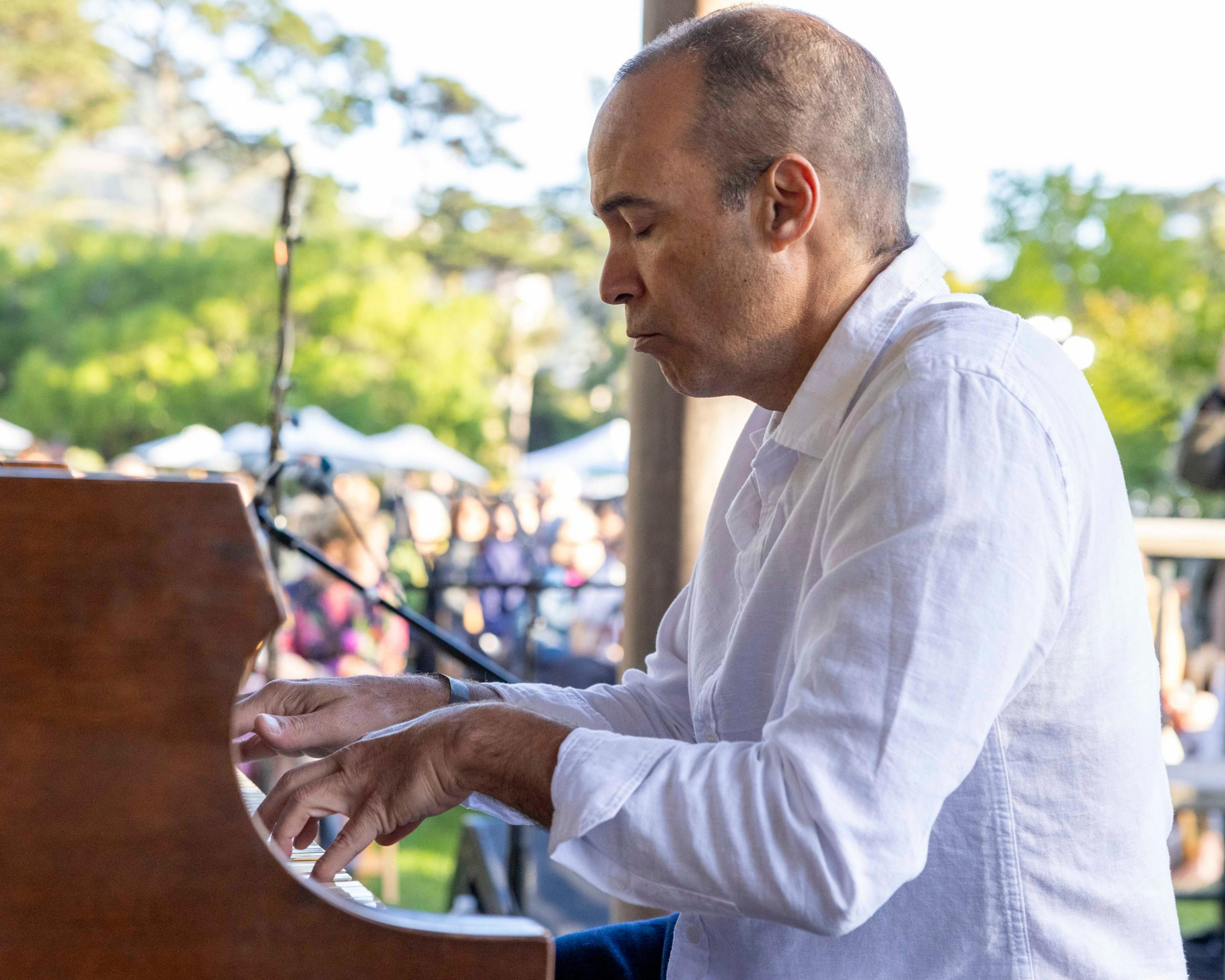 A man in a white shirt plays a piano outdoors, eyes closed, focused intently. In the background, there's a crowd of people and lush green trees.