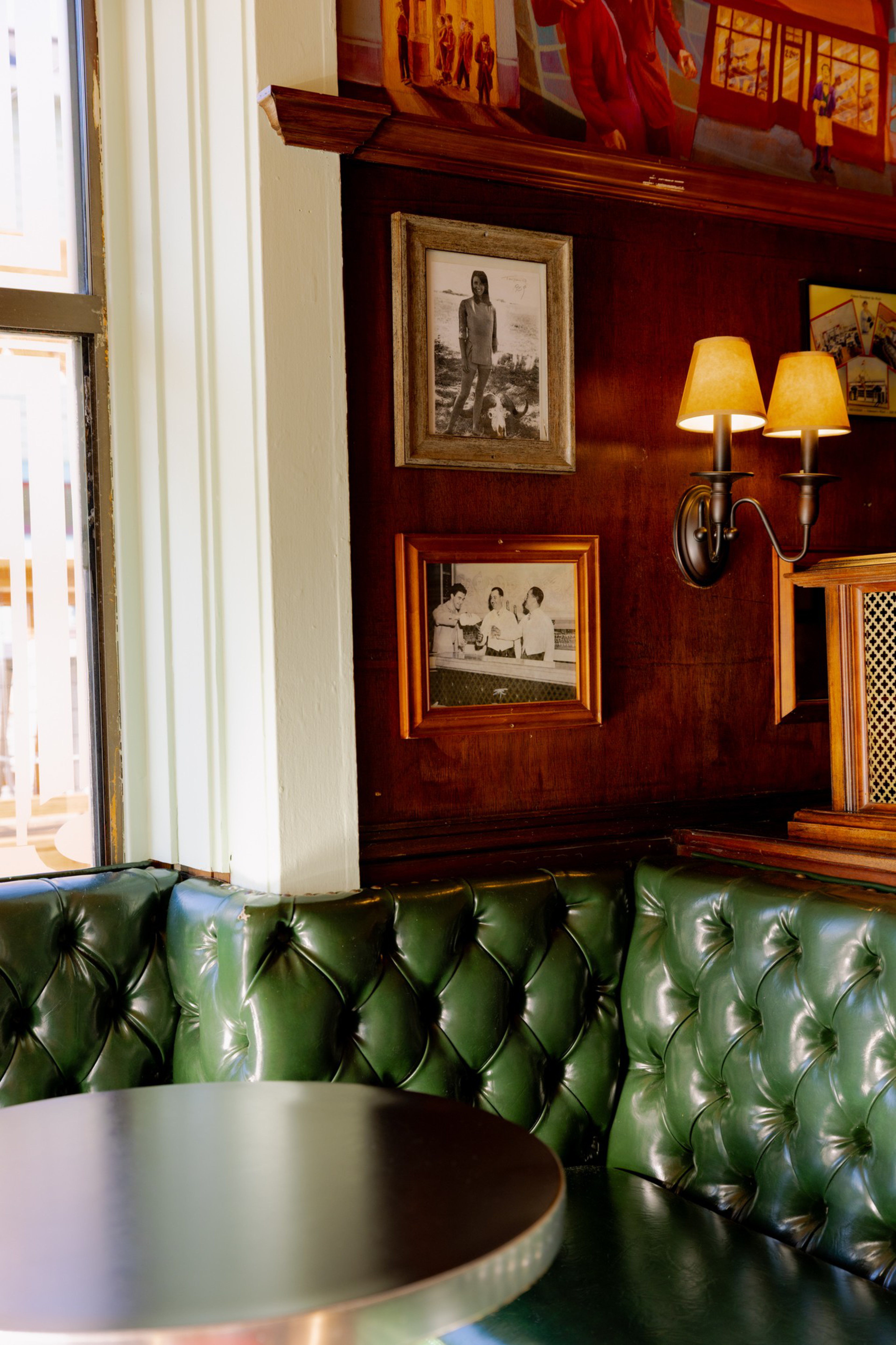 A green leather tufted booth and round table sit by a wood-paneled wall with black-and-white photos and a double-shaded wall sconce light.