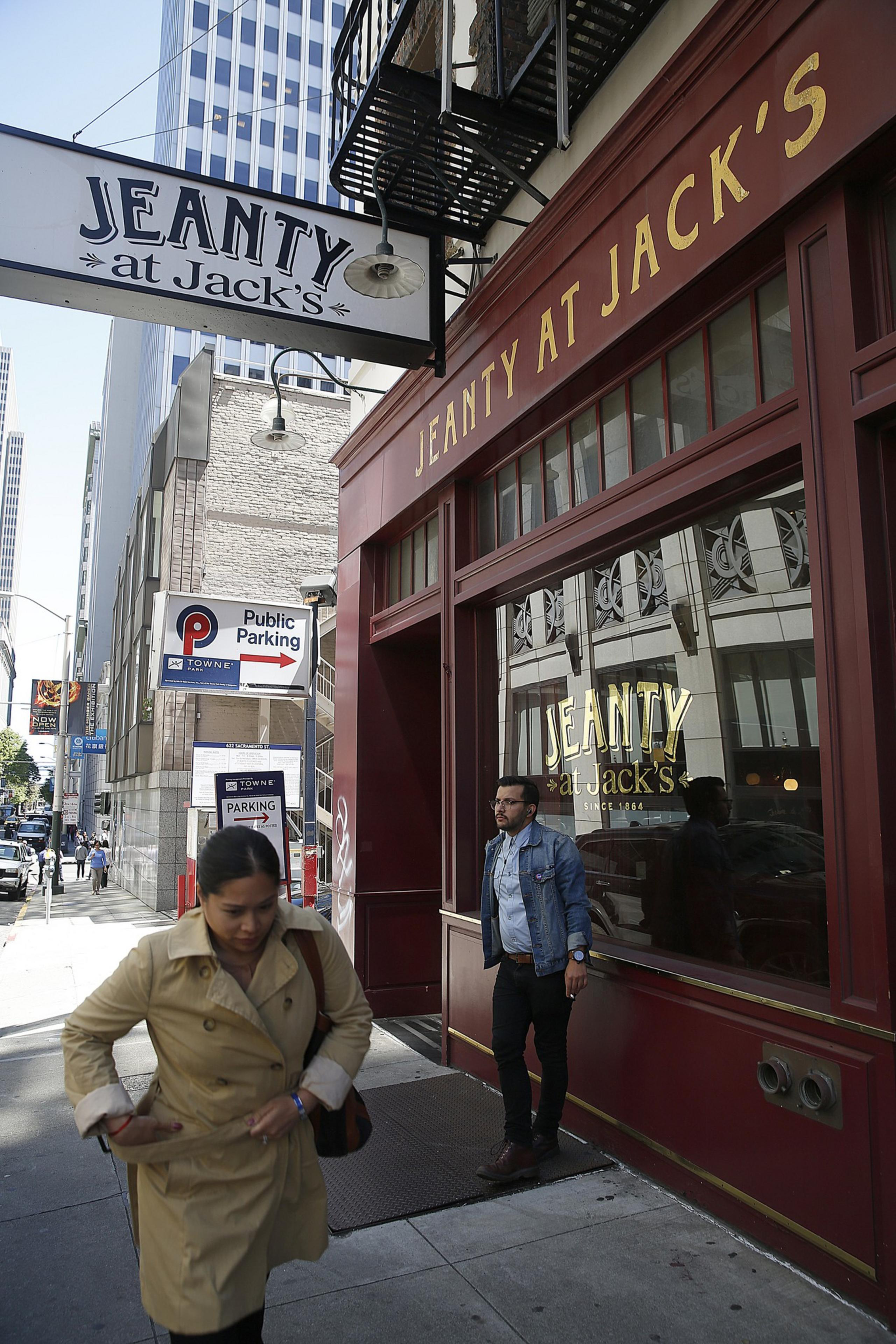 A woman in a beige trench coat walks past a man standing outside a red storefront with gold lettering "JEANTY AT JACK'S" on a city sidewalk.