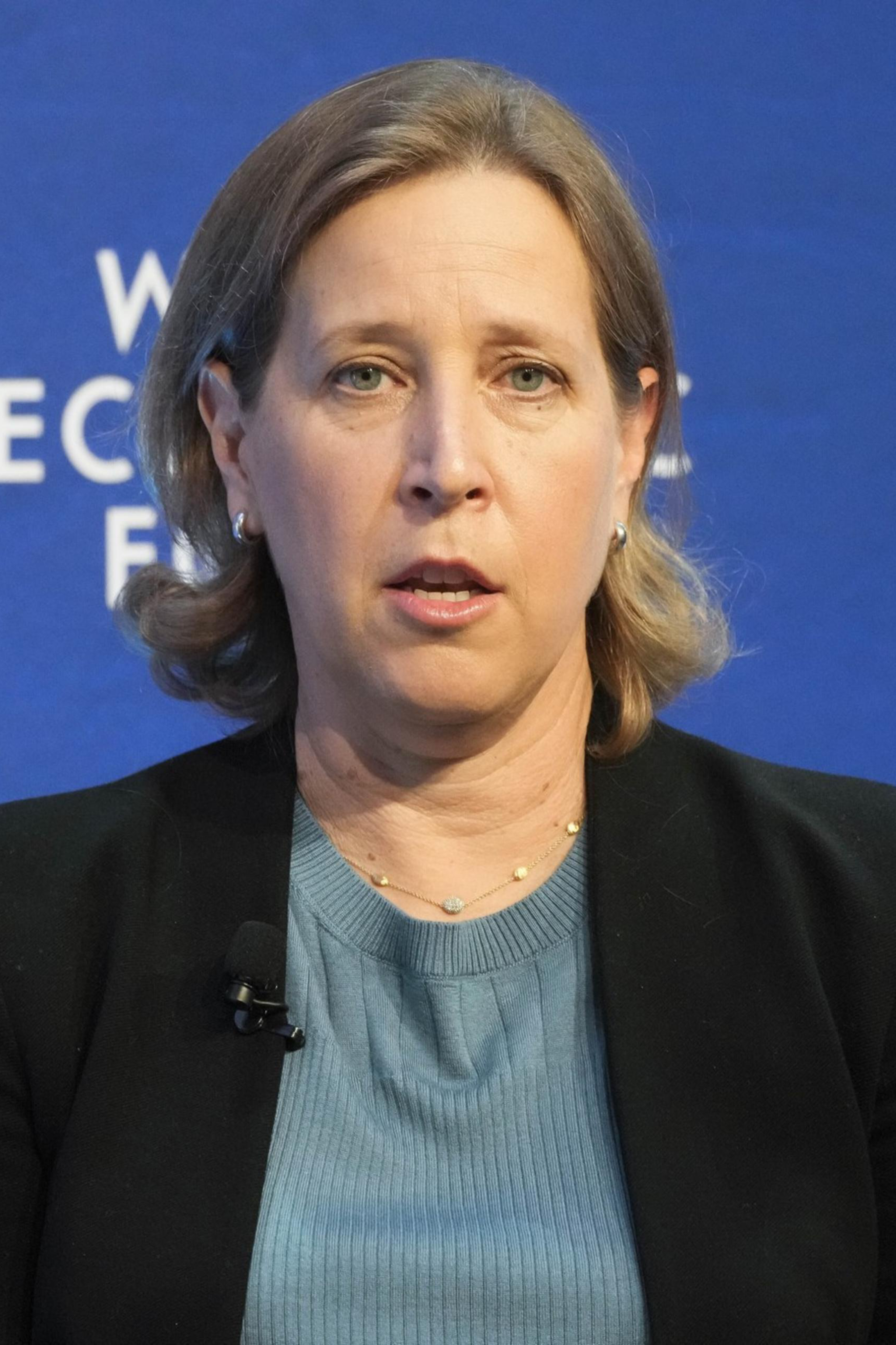 A woman with light brown hair speaks, raising one hand slightly. She is wearing a black blazer over a blue top. The background displays "World Economic Forum."