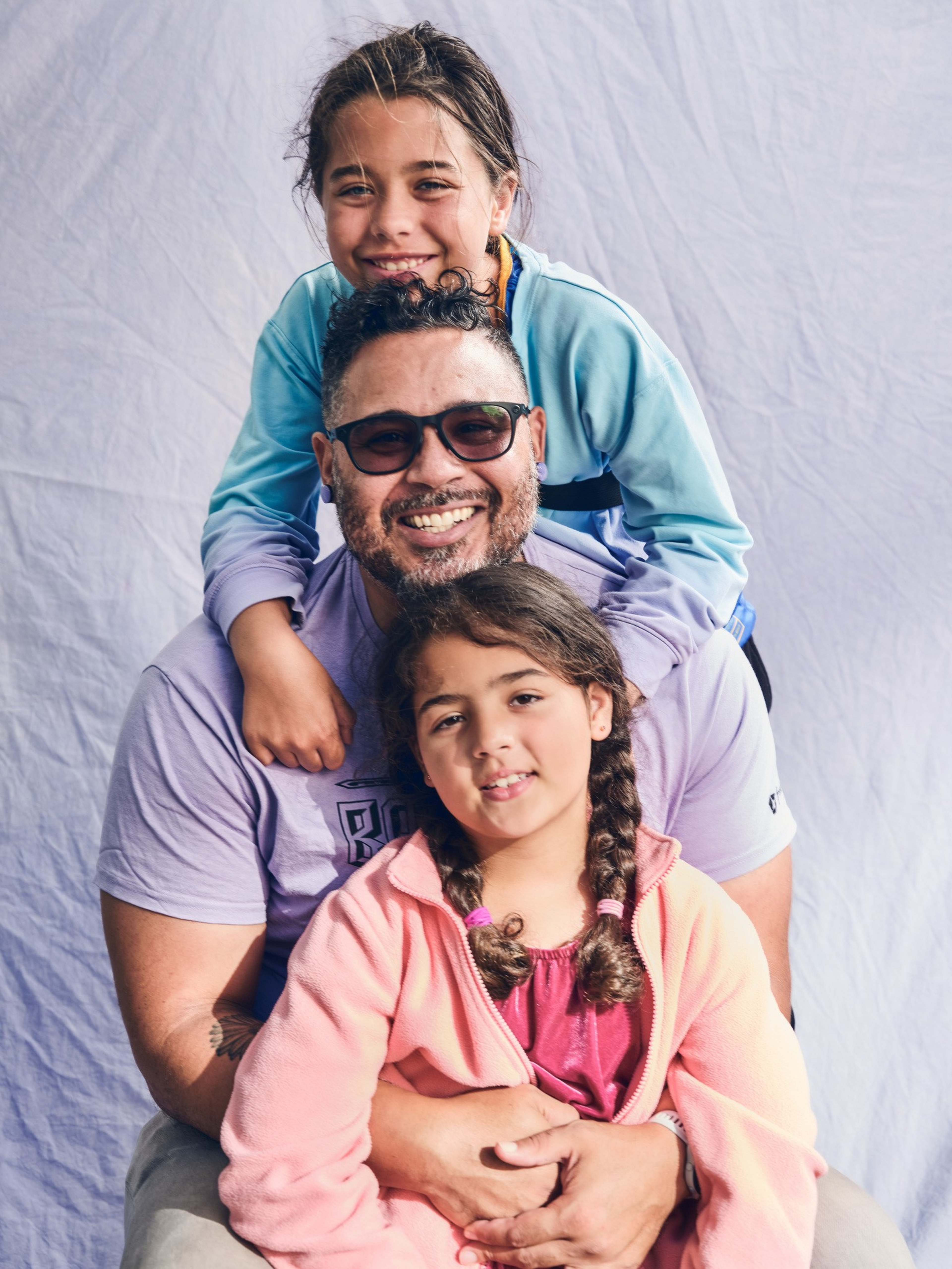 A man wearing sunglasses smiles while holding a young girl with braided hair; another girl in a blue jacket leans on his shoulders, all posing happily.