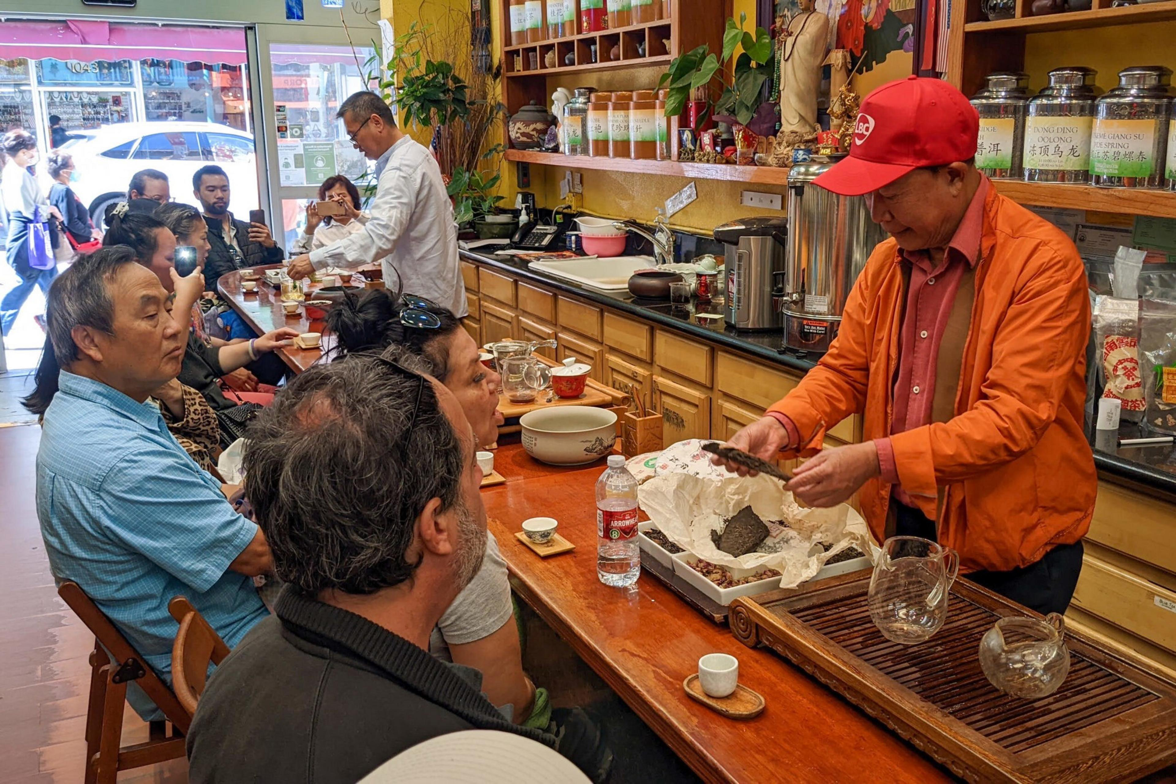 A man in an orange jacket and red cap prepares tea while several people sit at a wooden counter watching him in a cozy tea shop filled with jars and tea accessories.