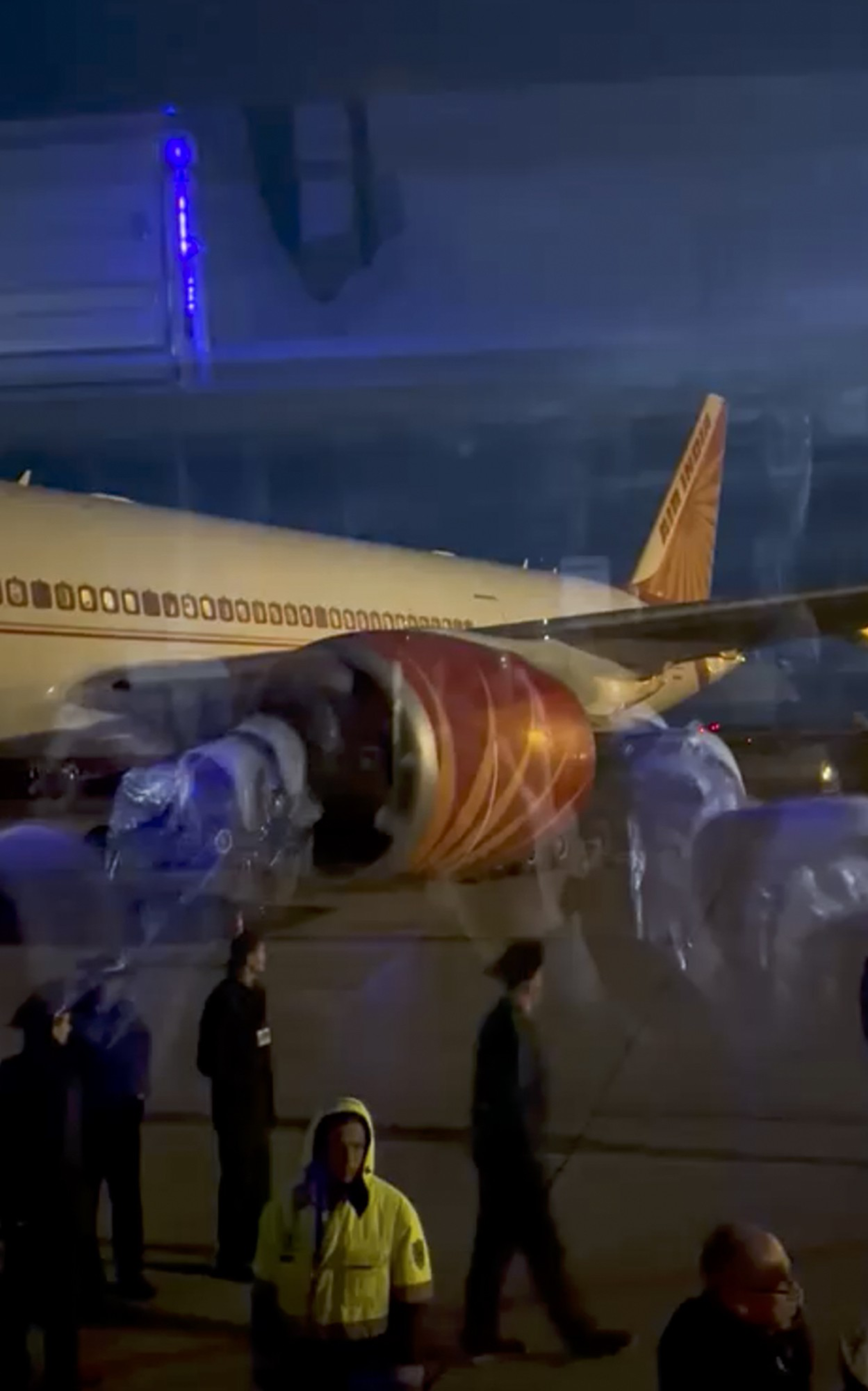 A large airplane with a red engine and Air India logo is parked on a nighttime tarmac, with several people standing nearby.