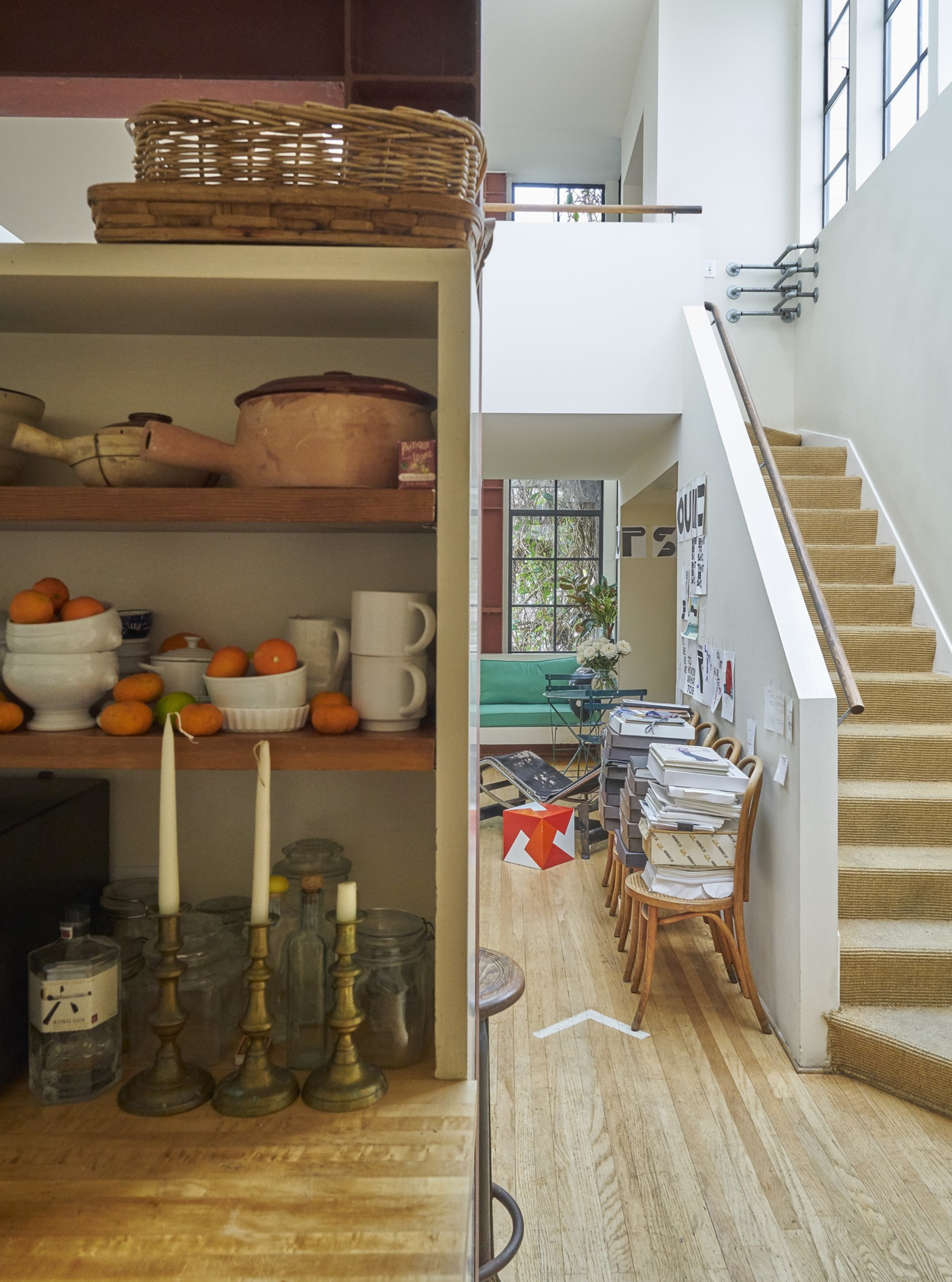 A cozy, eclectic home corner with a shelf of fruits and pottery, a staircase, a stack of books, chairs, and glimpses of a green sofa and art supplies.