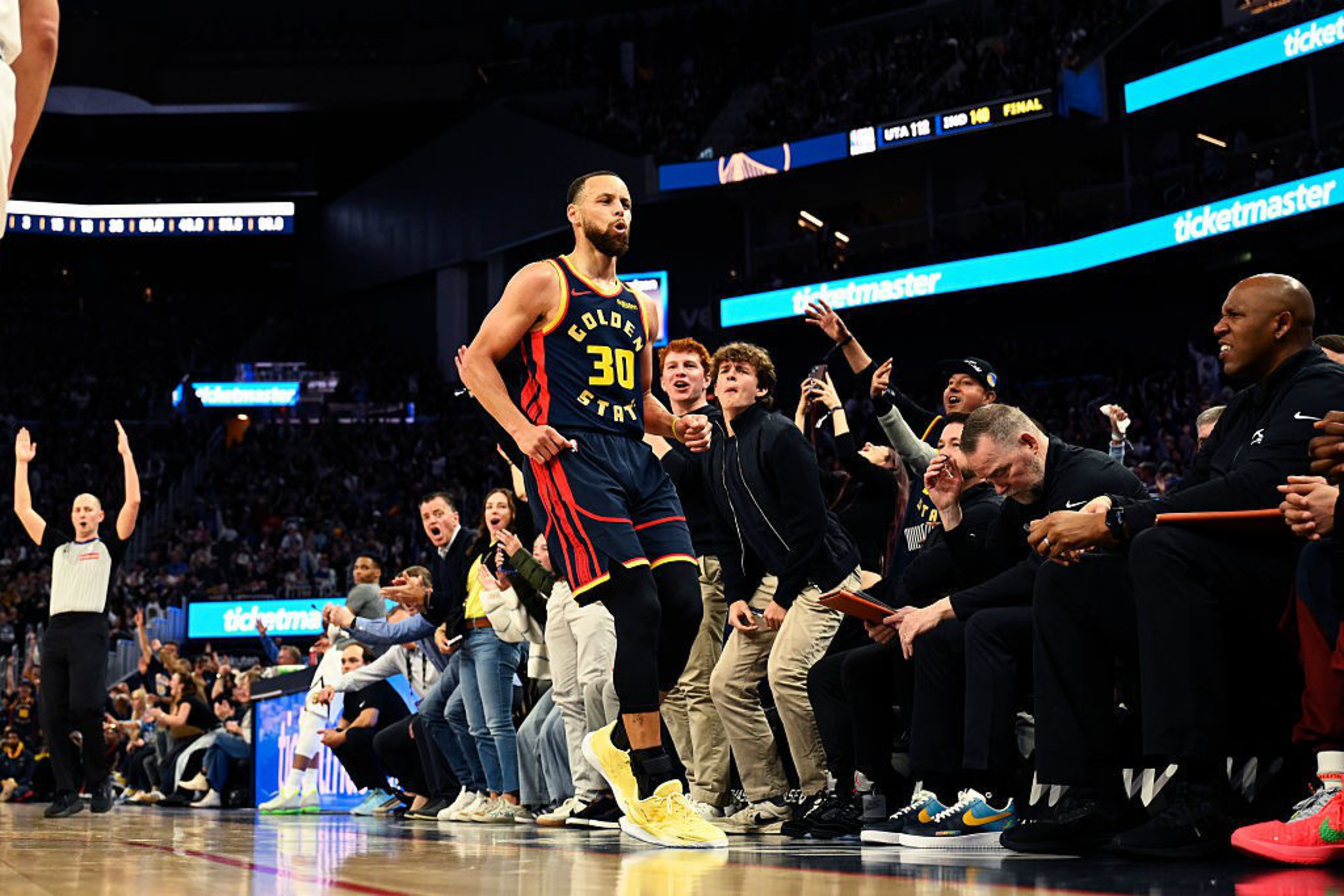 A basketball player in a Golden State jersey walks confidently near the sidelines as the crowd around him stands and cheers energetically.