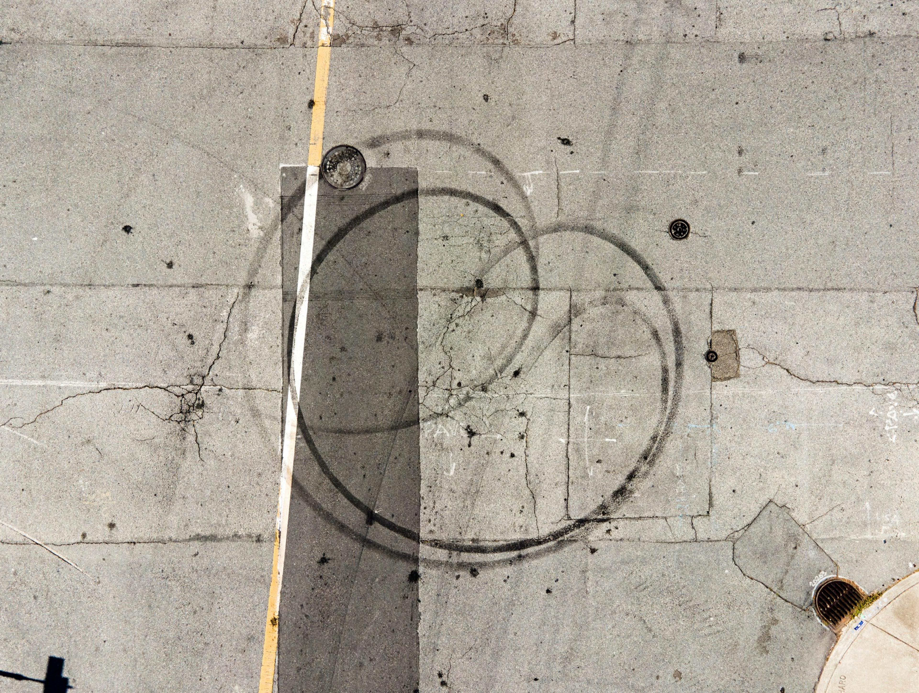 Aerial view of a cracked asphalt surface with circular tire marks, a yellow line, and two manholes, one on the left and another on the right near a drain.