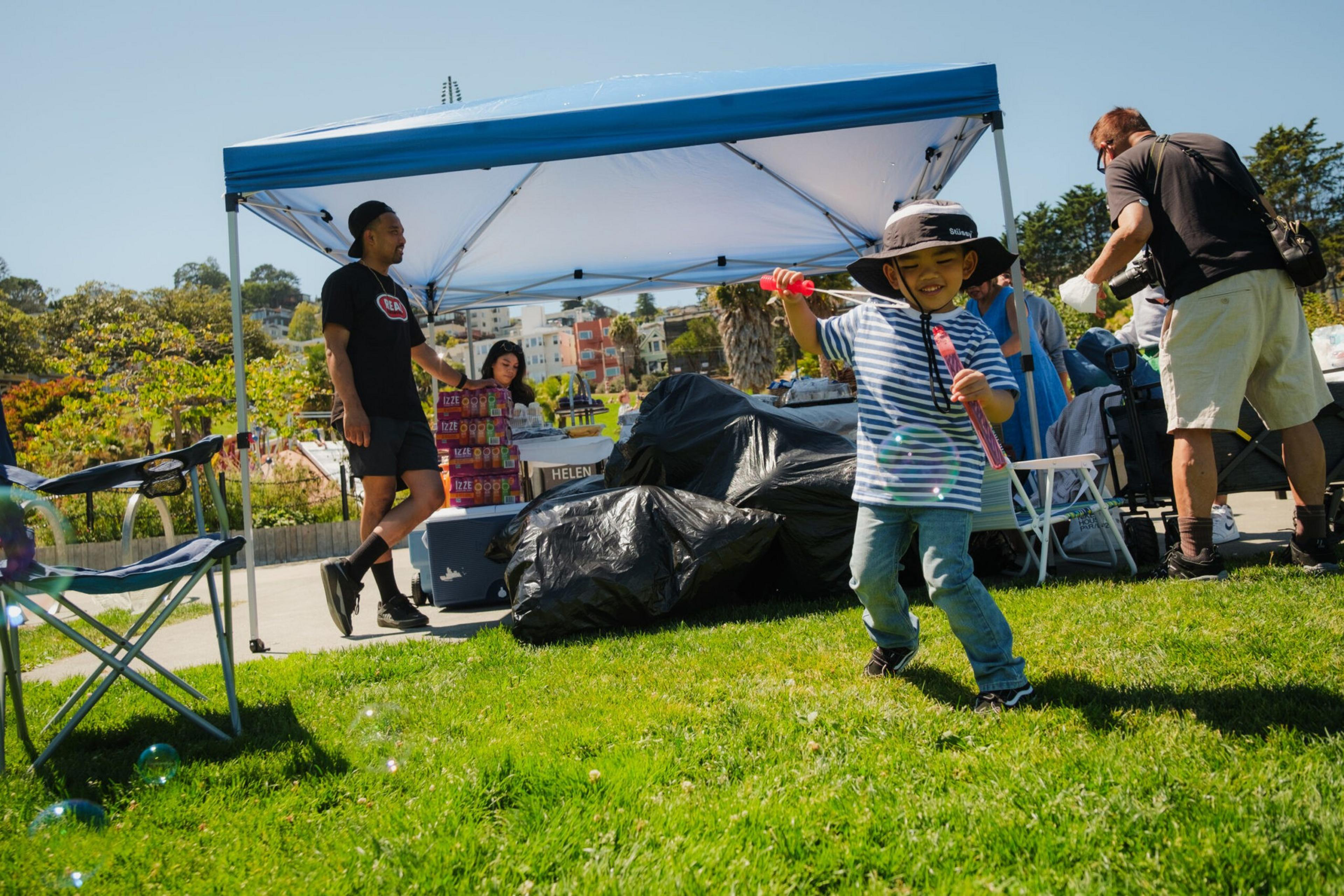 A child in a striped shirt and sun hat happily blows bubbles on green grass near a blue canopy where adults stand and sit with supplies.