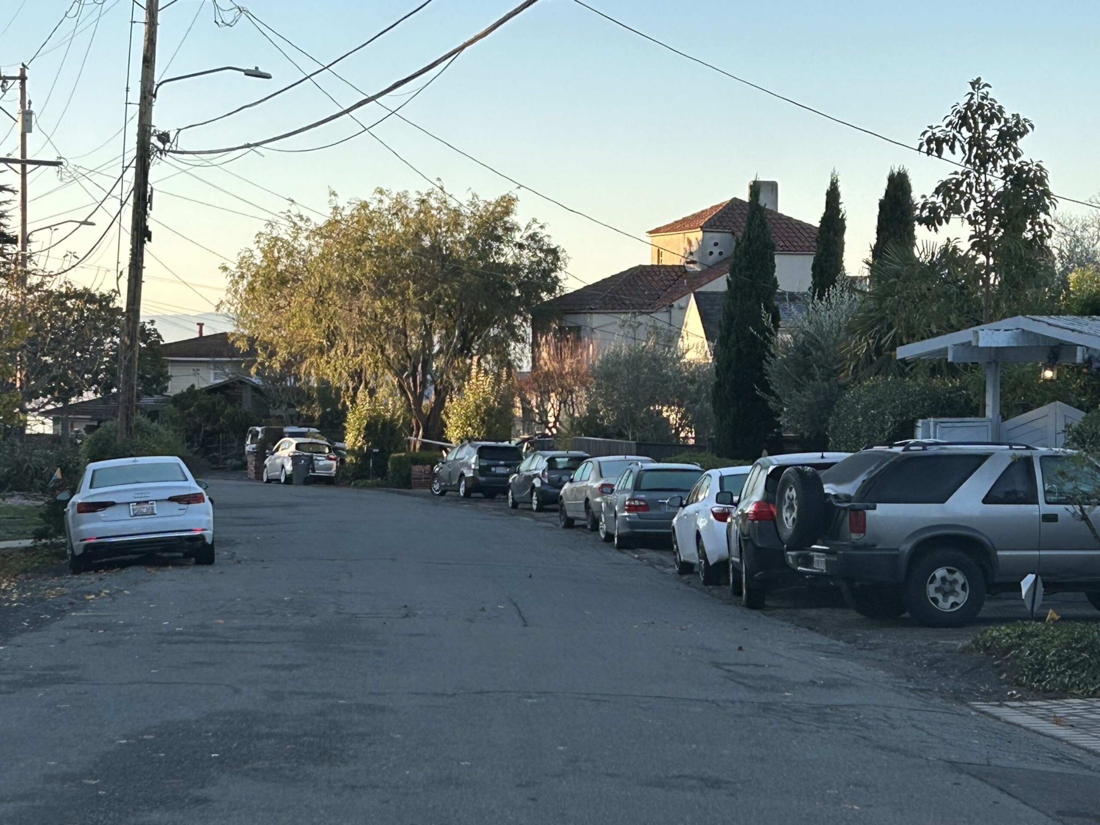 A quiet residential street lined with parked cars on both sides, trees, power lines overhead, and houses with tiled roofs in the background.
