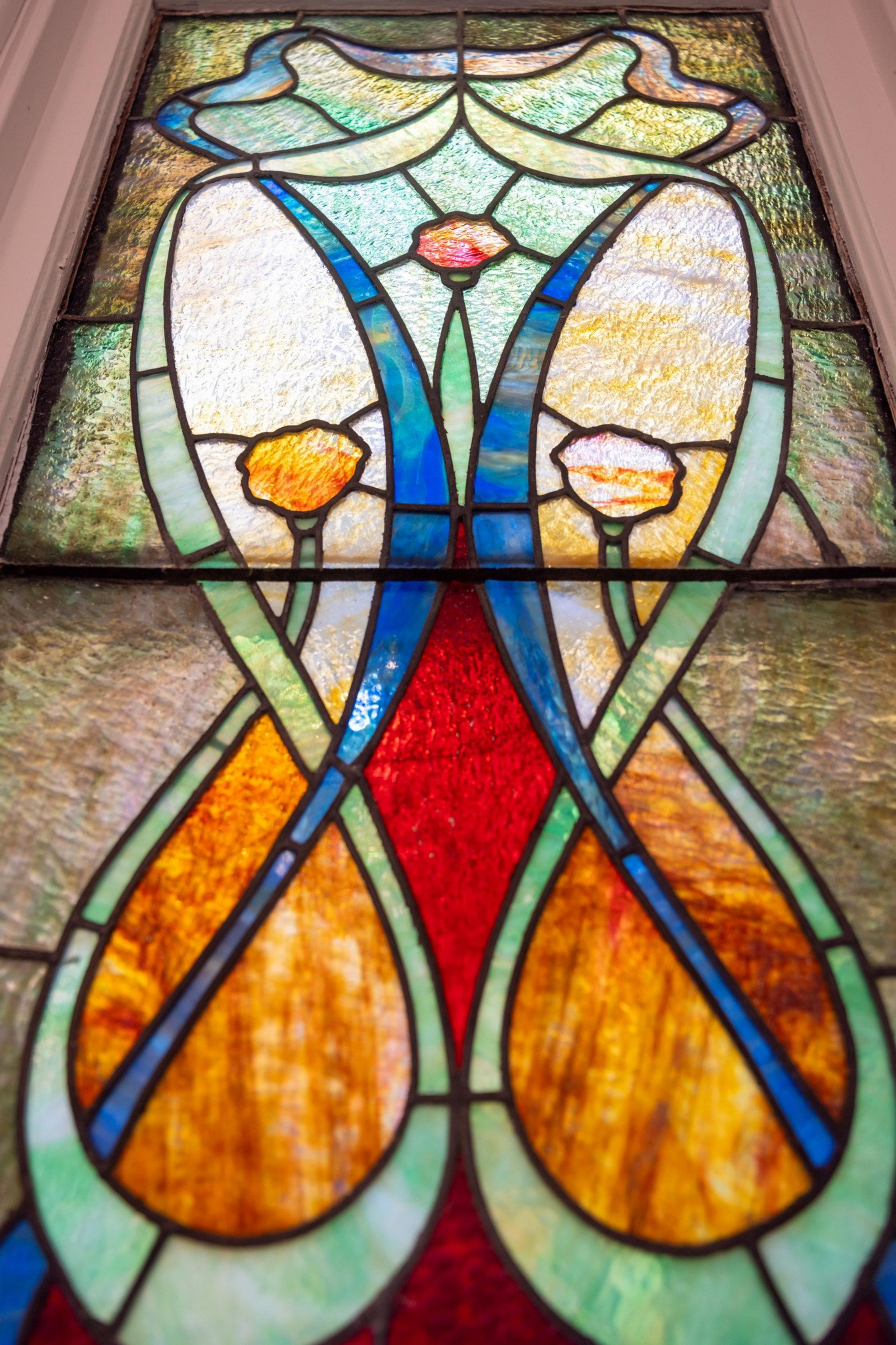 This image shows a colorful stained-glass window with an intricate floral design in shades of red, yellow, green, and blue, set in a wooden frame.