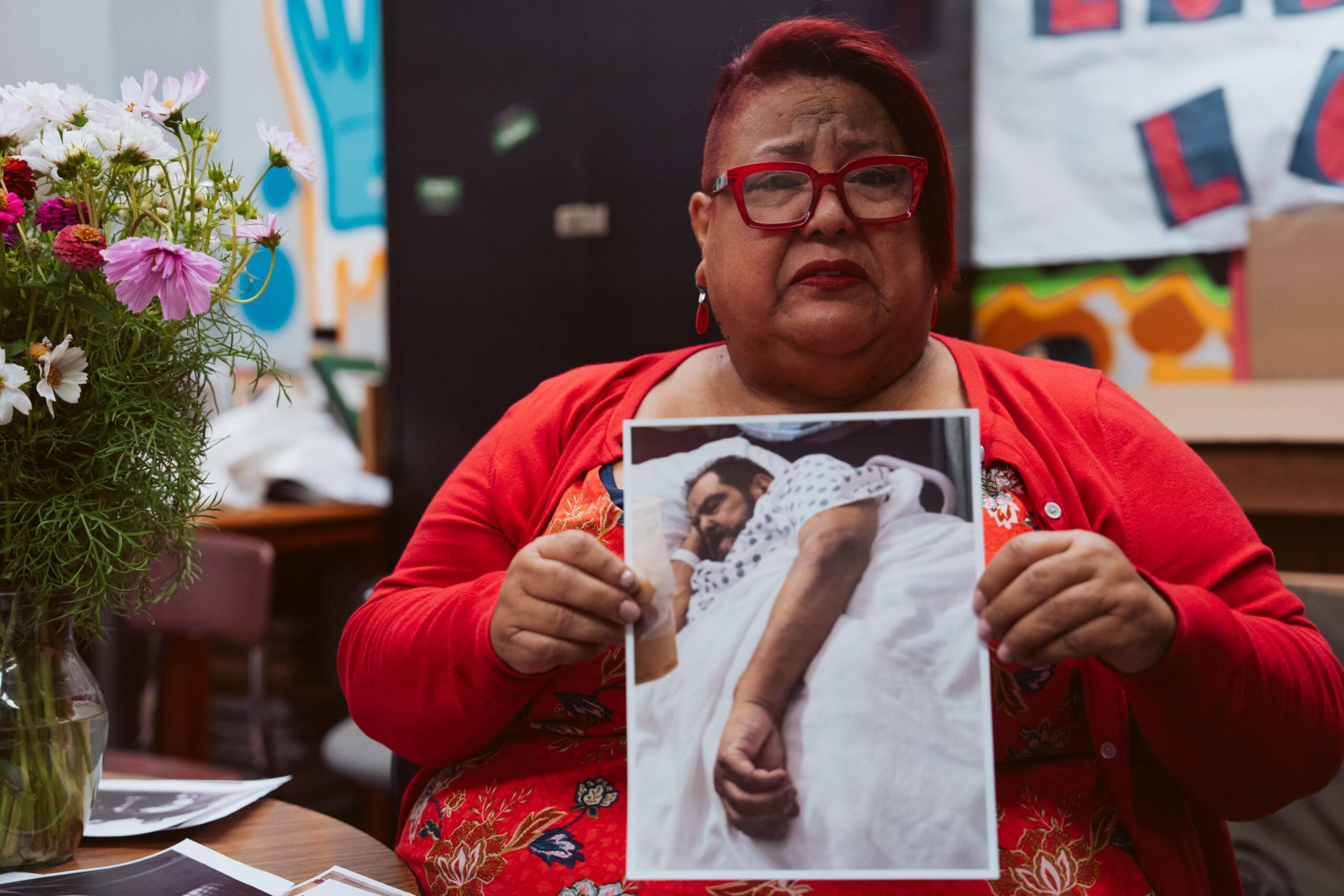 A woman with red glasses and clothing holds a photo of a man lying in a hospital bed wearing a hospital gown.