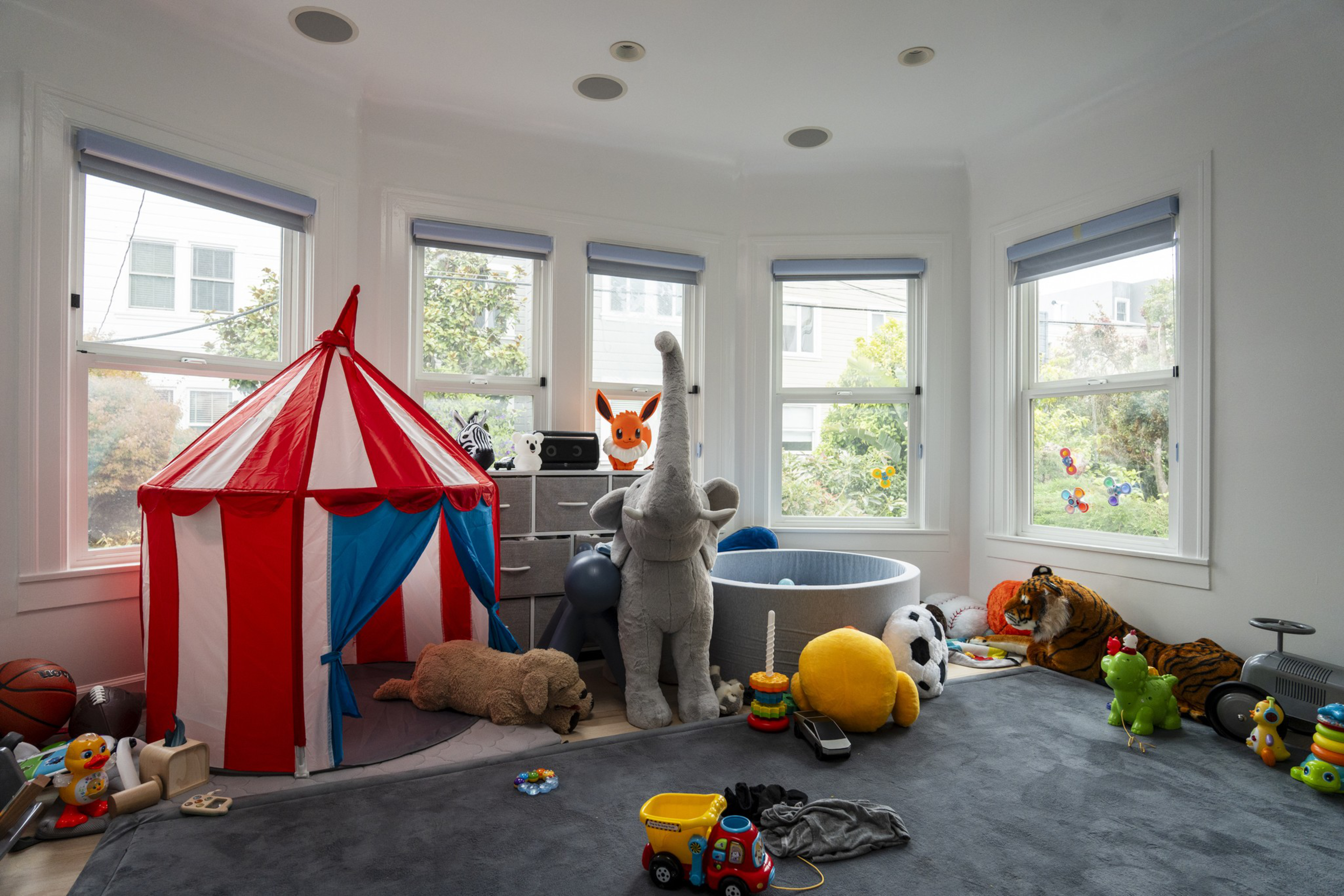 A bright playroom with large windows contains a red-and-white striped play tent, numerous stuffed animals, toys, and a small ball pit on a gray carpet.