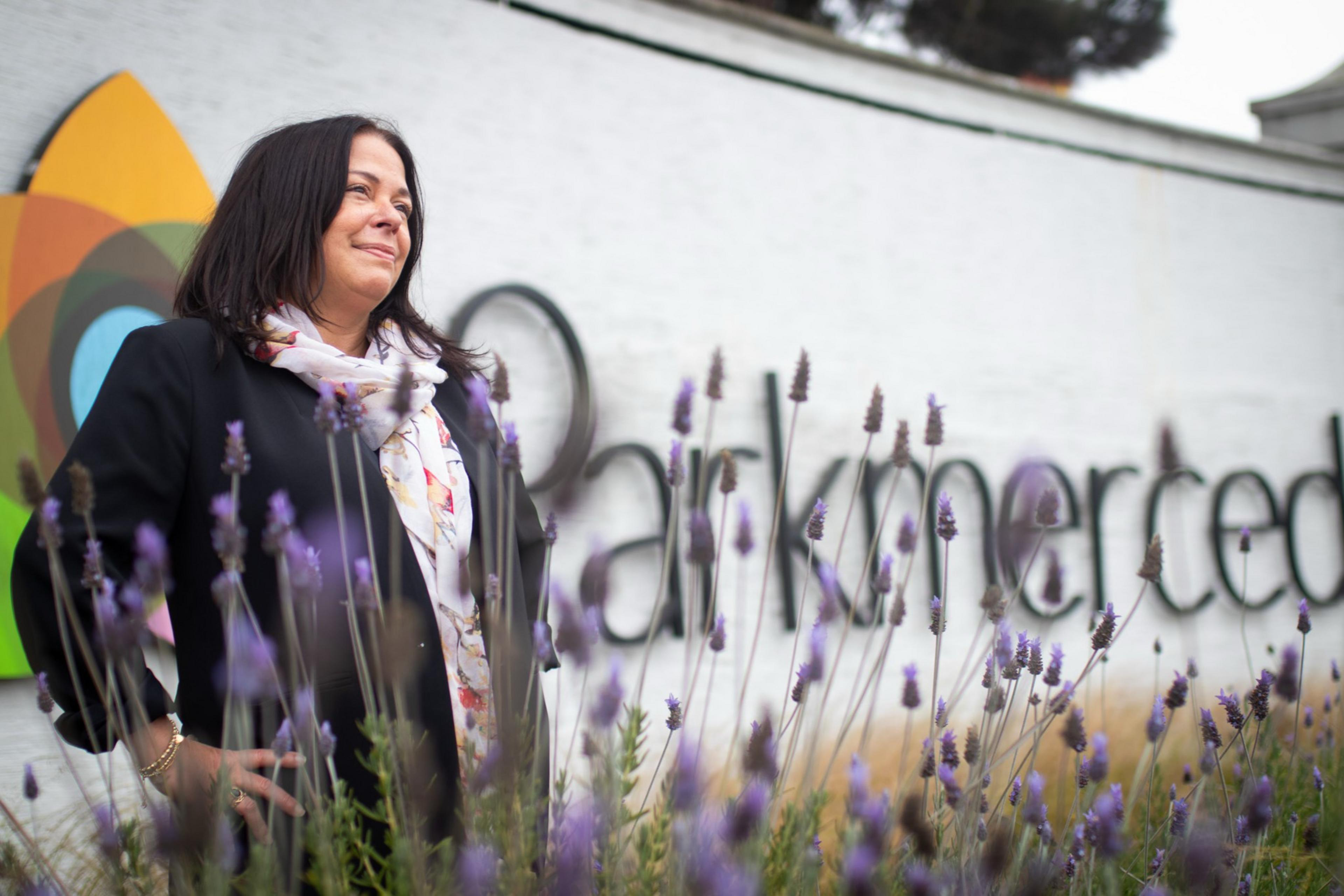 A woman stands confidently among lavender flowers. She is wearing a black jacket and floral scarf. Behind her is a partially visible sign reading "Parkmerced."