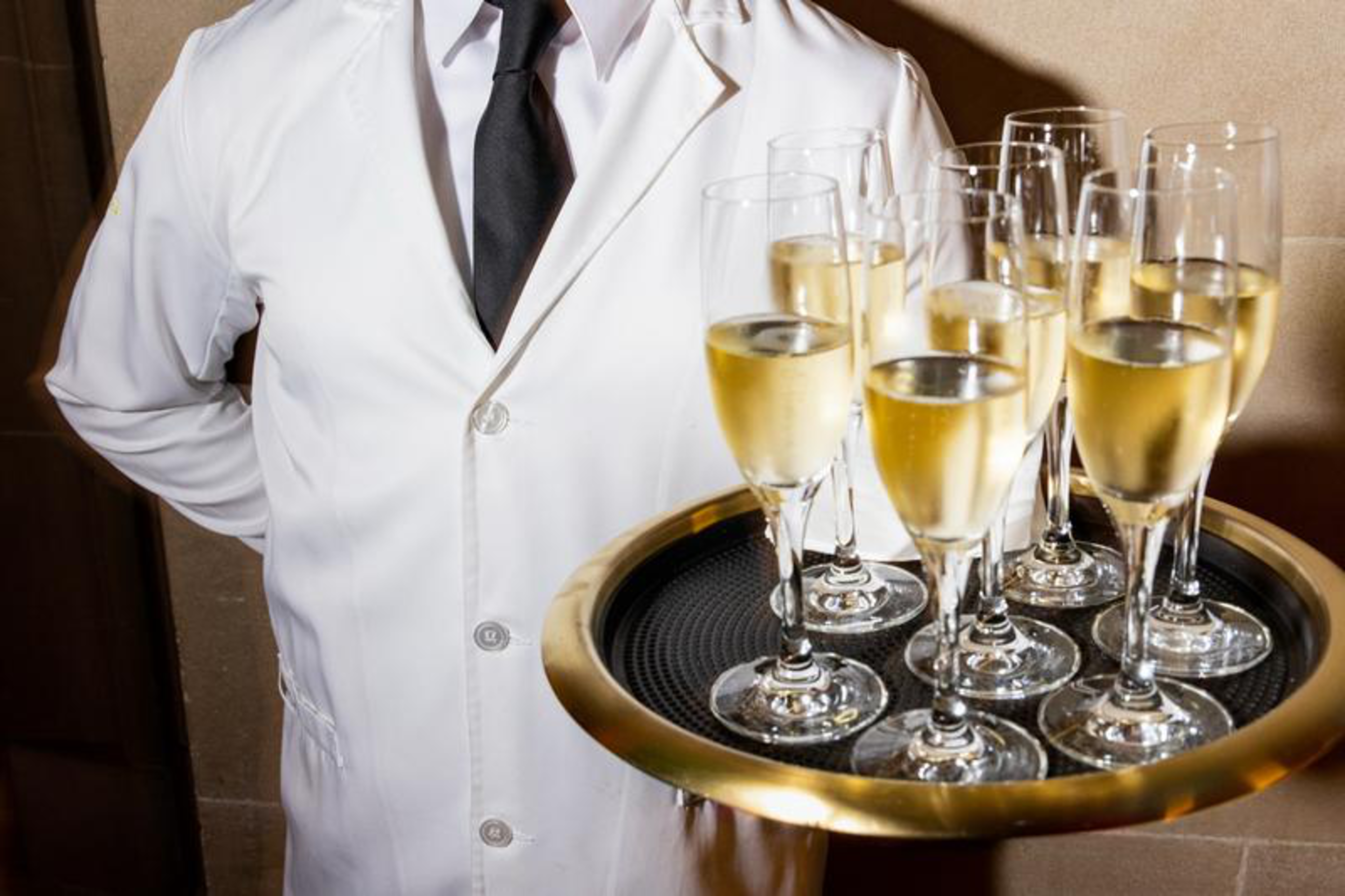 A waiter dressed in a white coat and black tie holds a round tray with eight filled champagne glasses.