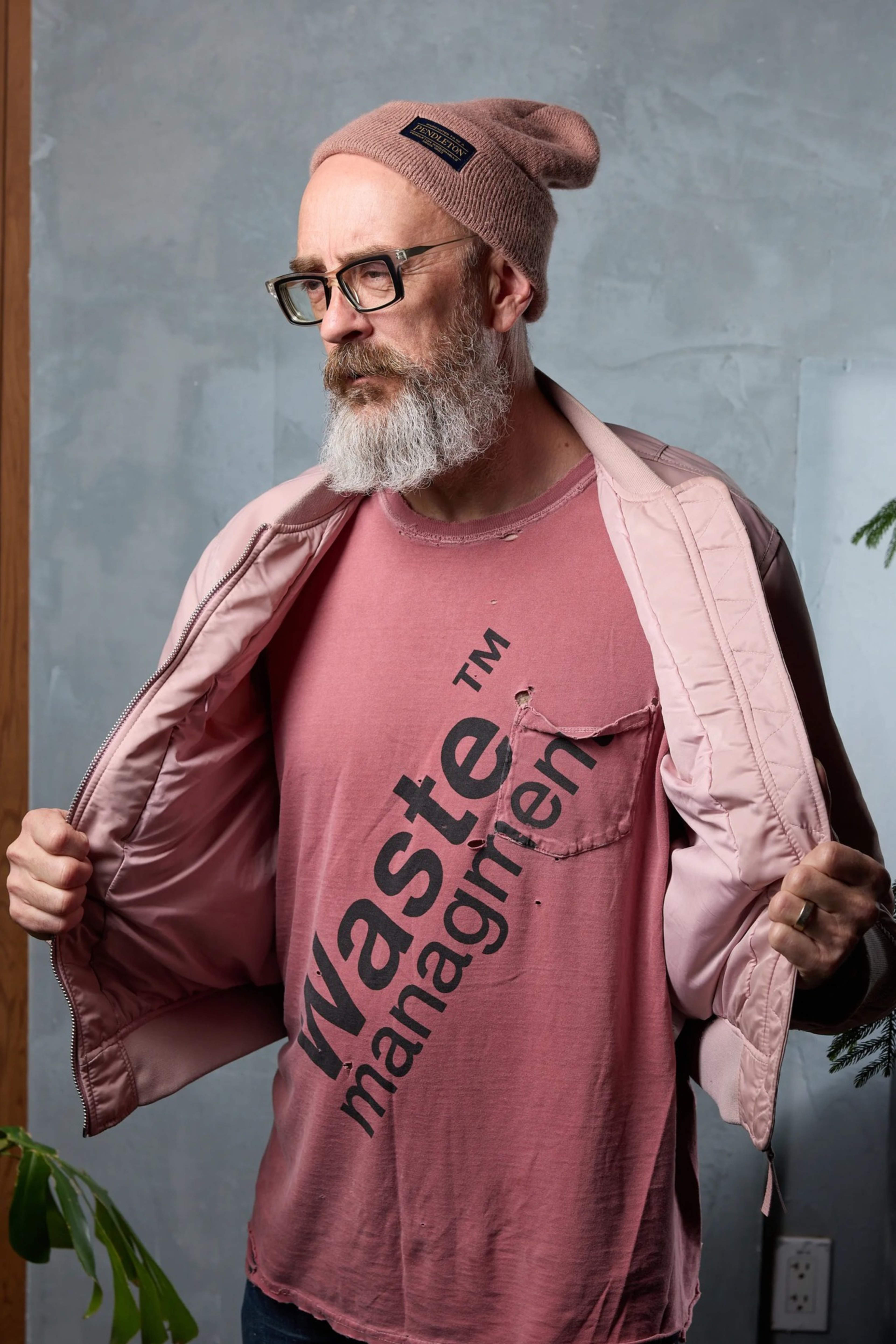 A man with a beard and glasses wears a pink beanie and t-shirt reading &quot;waste management.&quot; He holds open a pink jacket with a serious expression.