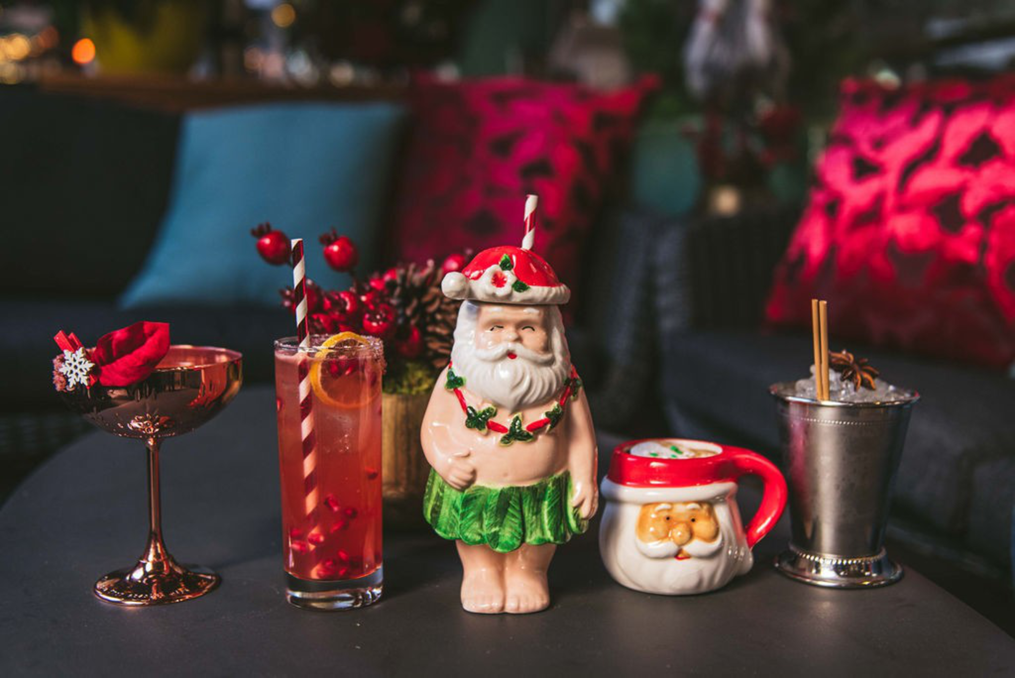 A lineup of holiday-themed cocktails, including drinks in brass martini glass, a Santa-shaped mug, and a cup with Santa wearing a hula skirt.