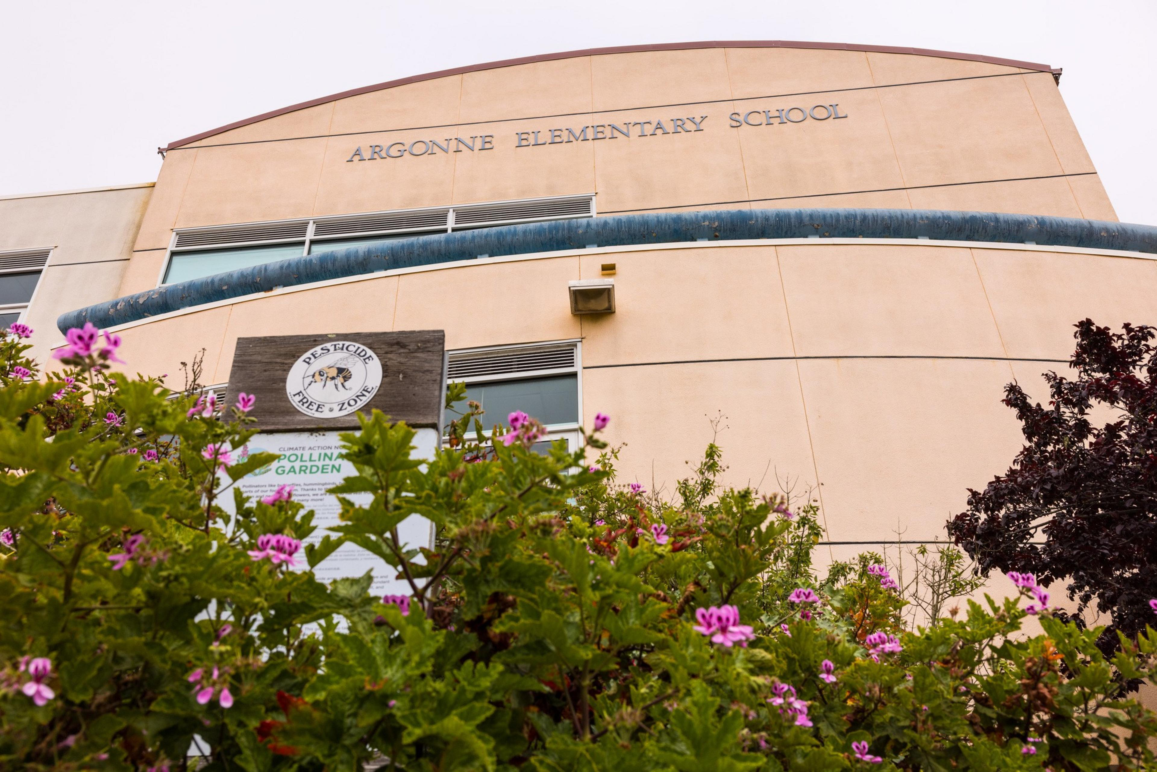 The image shows Argonne Elementary School's curved facade, with "Pesticide Free Zone" and garden signs, framed by blooming pink flowers and greenery.