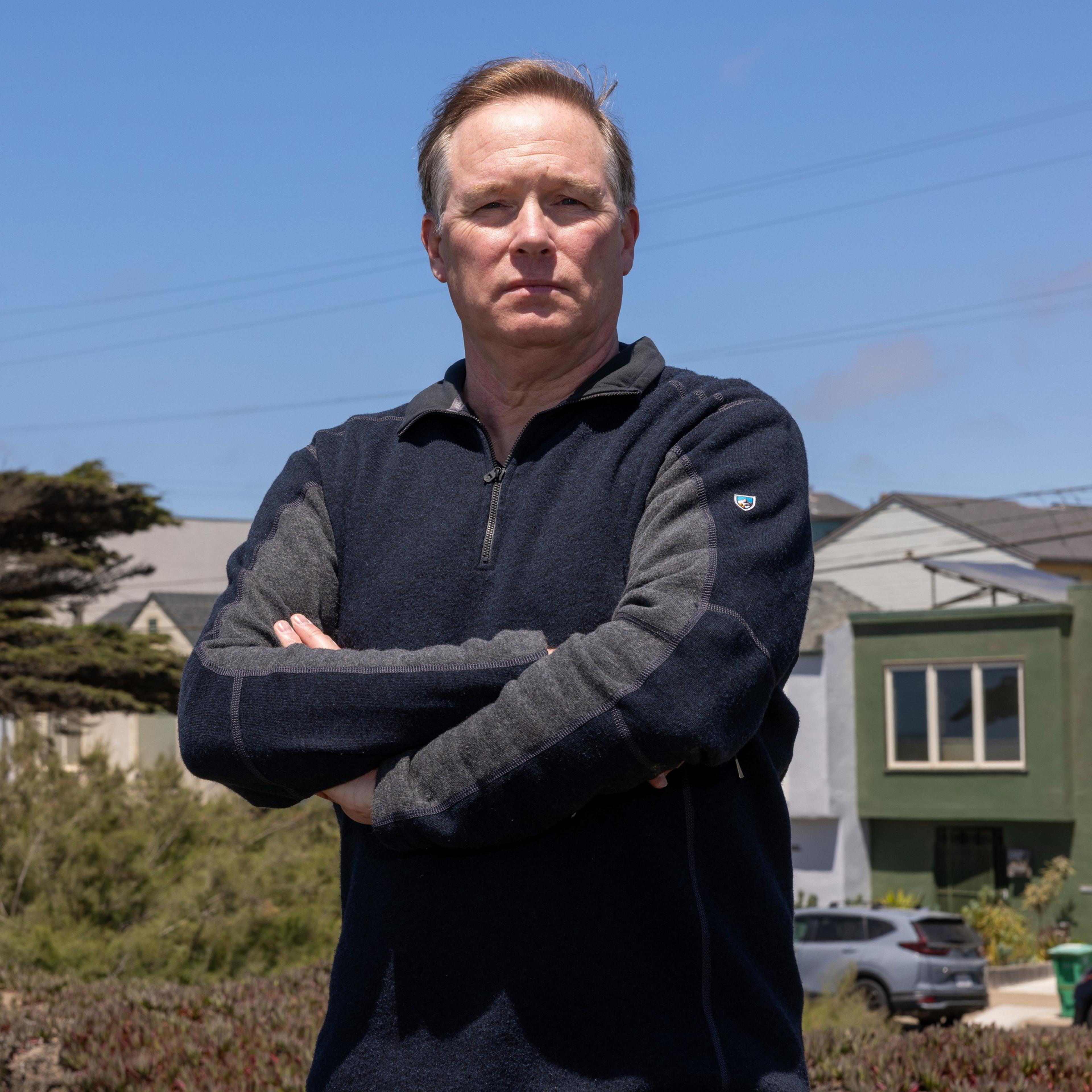 A man is standing with arms crossed, wearing a dark long-sleeve shirt. He's outside with a blue sky, houses, and greenery in the background. His expression is serious.