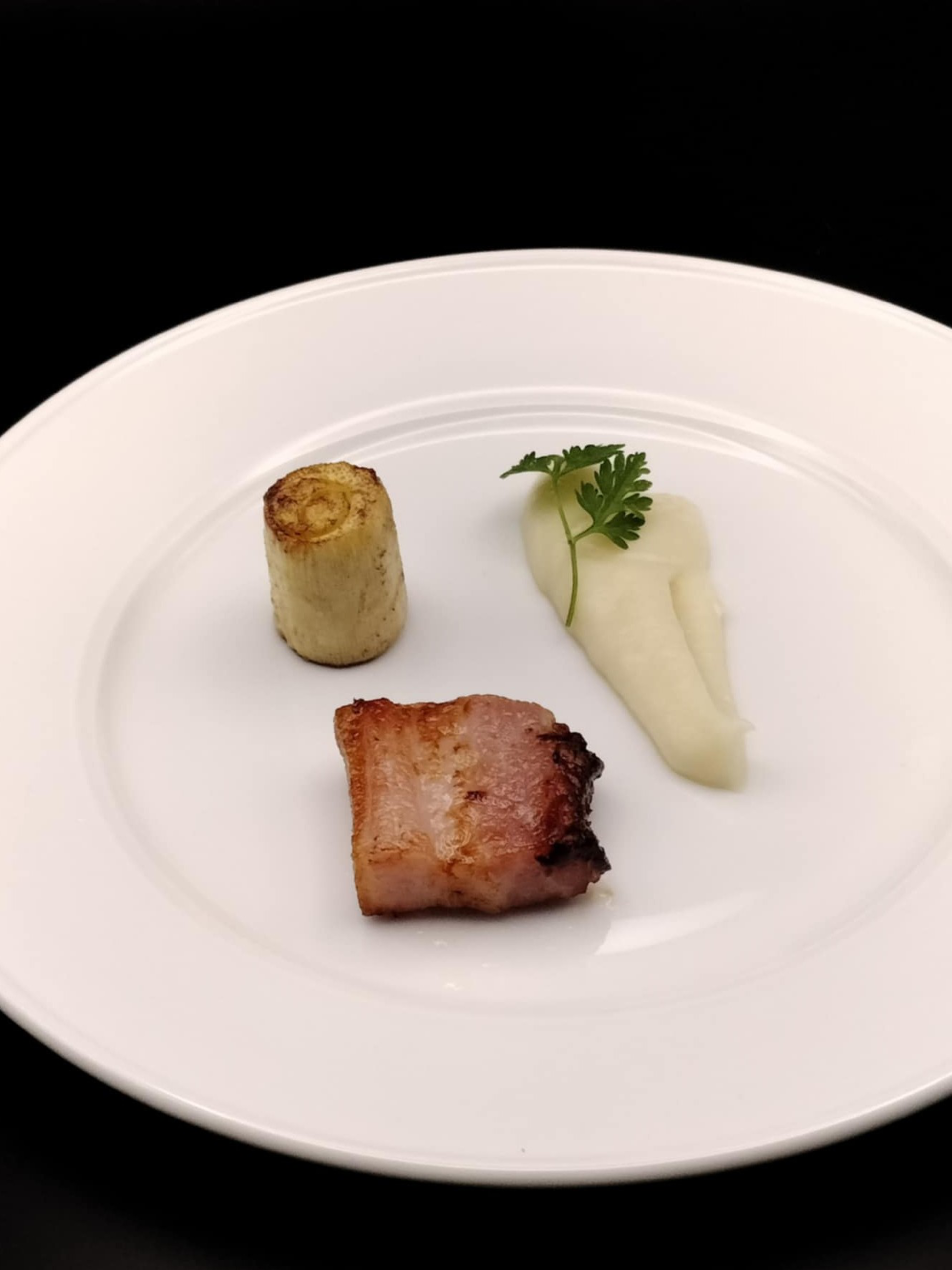 A small, seared piece of meat, a cylindrical roasted vegetable, and a dollop of creamy puree garnished with a green herb leaf on a white plate.