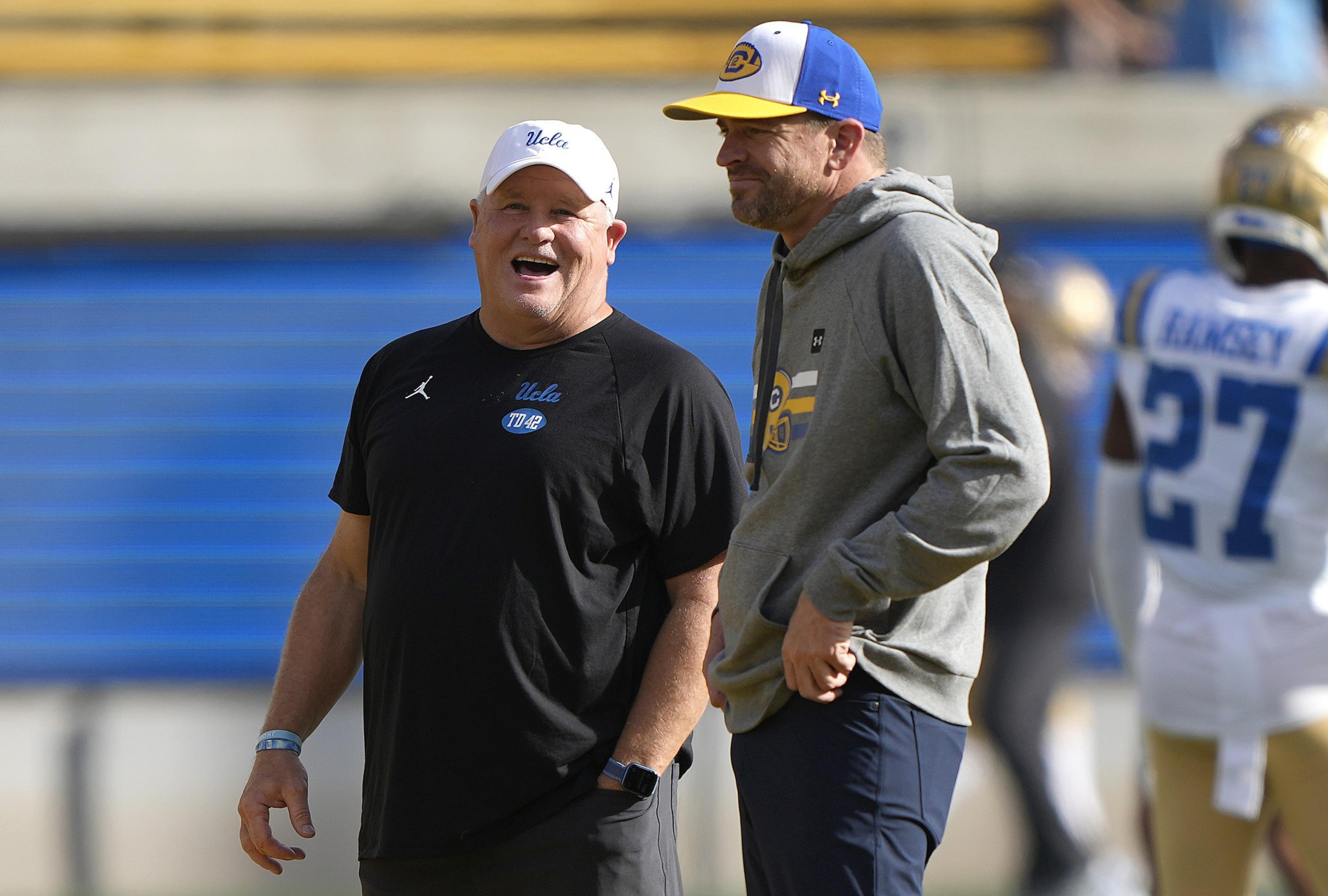 A man in a black UCLA shirt and white cap laughs next to a man in a gray hoodie and blue-yellow cap, with a football player in the background.