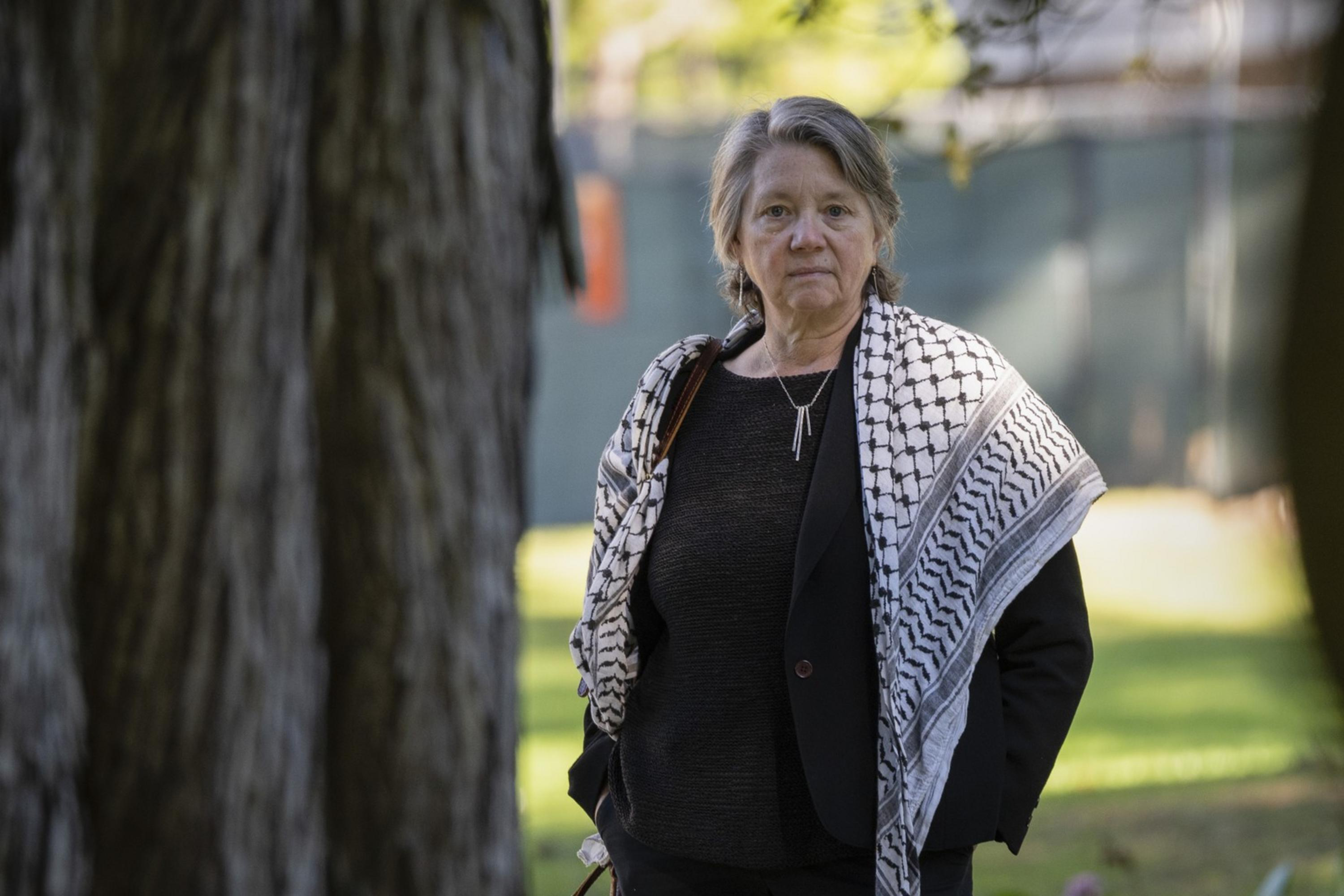 A person stands in a park, wearing a dark outfit and a patterned scarf. They have short, gray hair and the background is filled with trees and greenery.