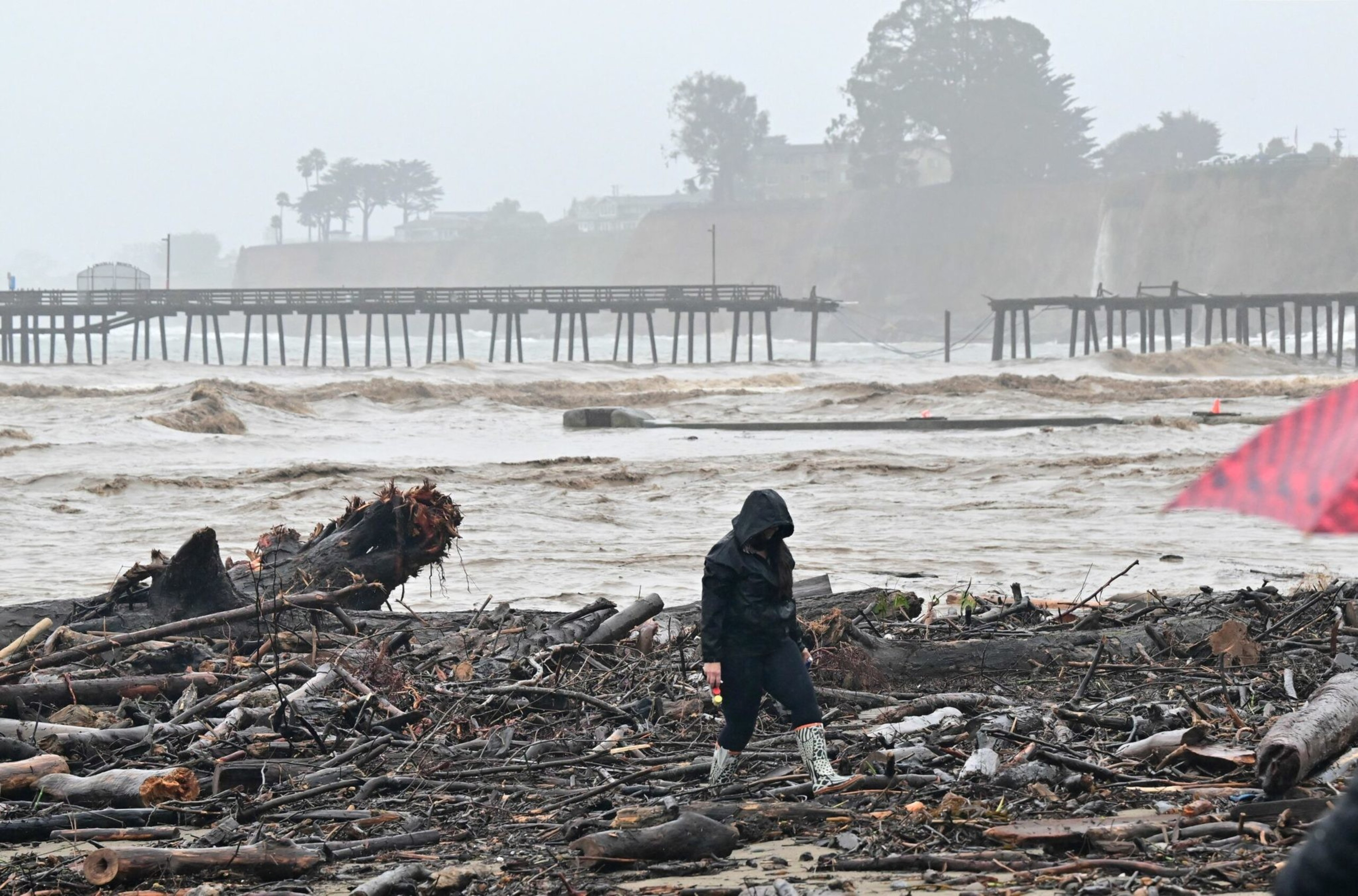 A person in rain gear walks through debris and driftwood on a muddy beach with a broken pier and choppy water in the background under gray skies.
