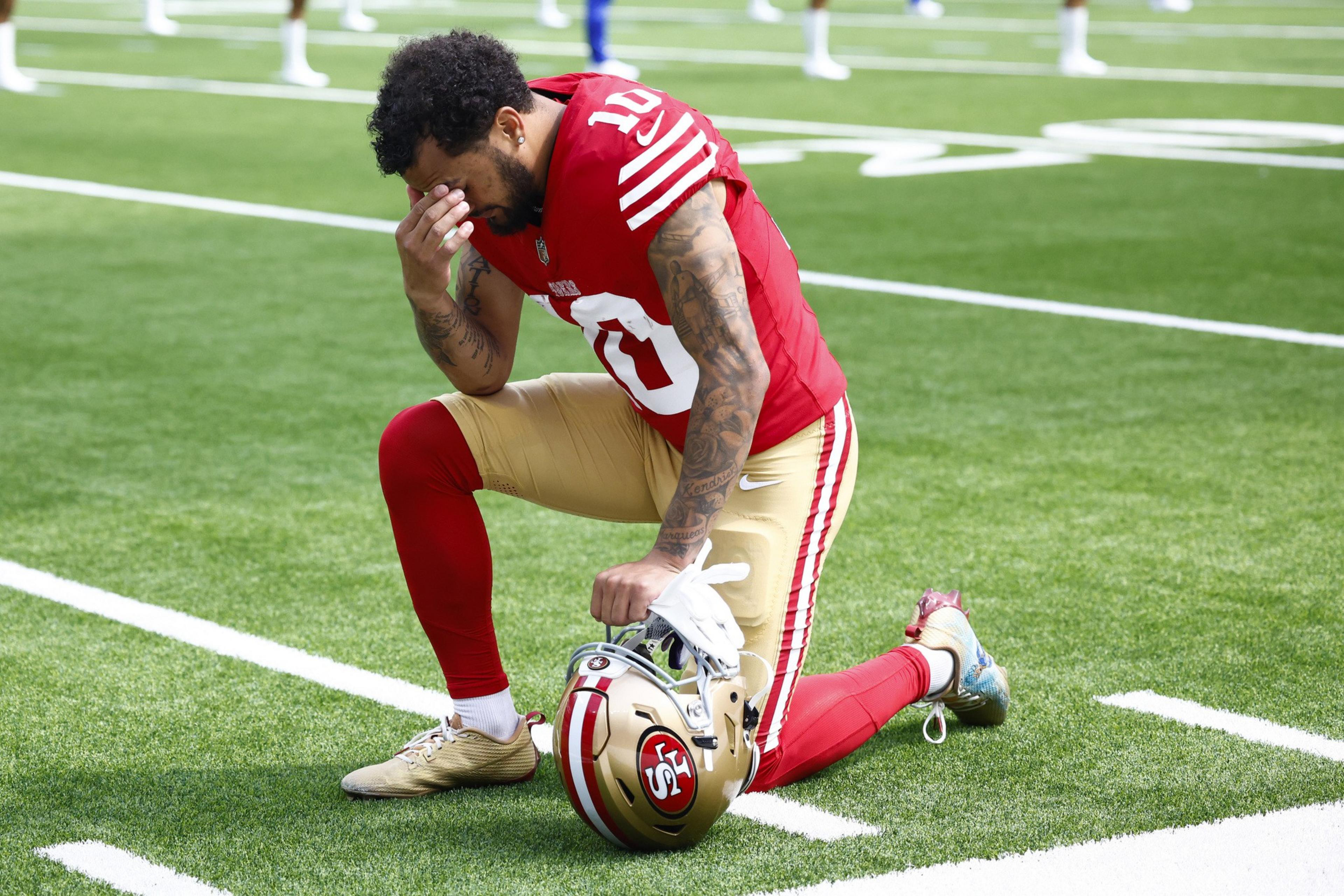 A football player in red and gold uniform kneels on a field, holding his helmet and touching his face as if in contemplation or prayer.