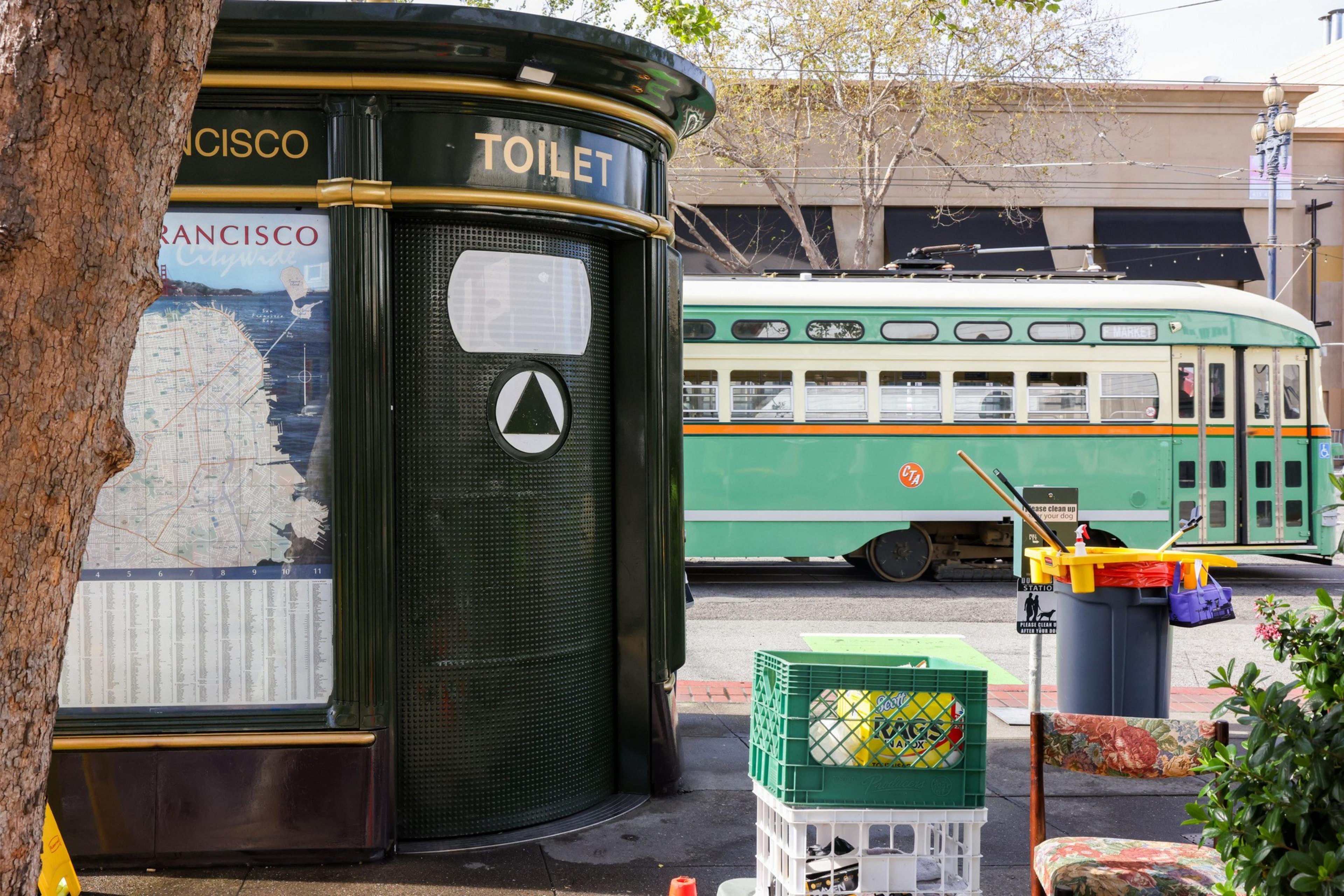 A green public toilet with a city map on it is next to a tree. Behind it, an old green streetcar is parked. Cleaning supplies and a crate are nearby.