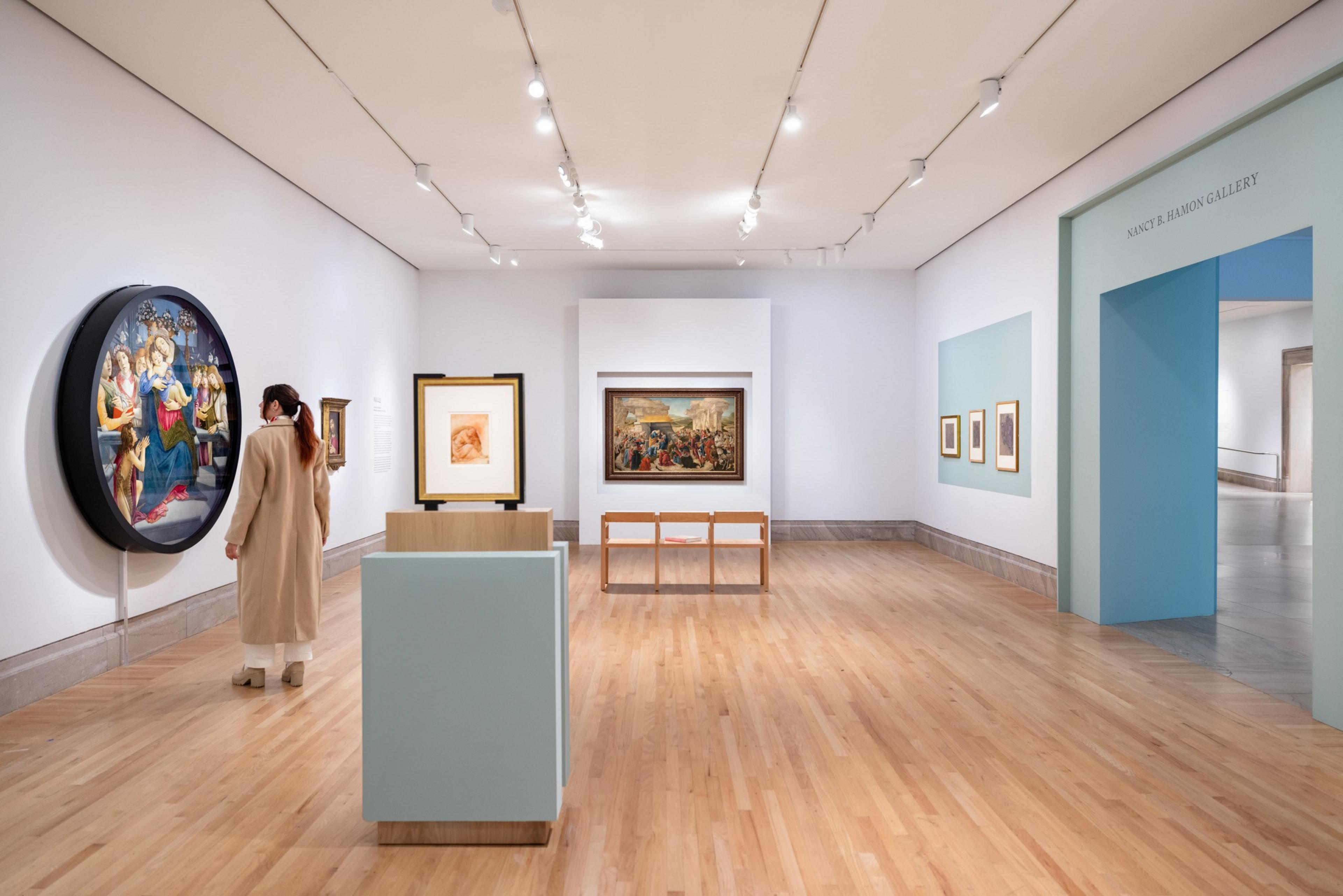 A woman in a long beige coat looks at a round oil painting in the exhibition Botticelli Drawings at San Francisco's Legion of Honor museum.