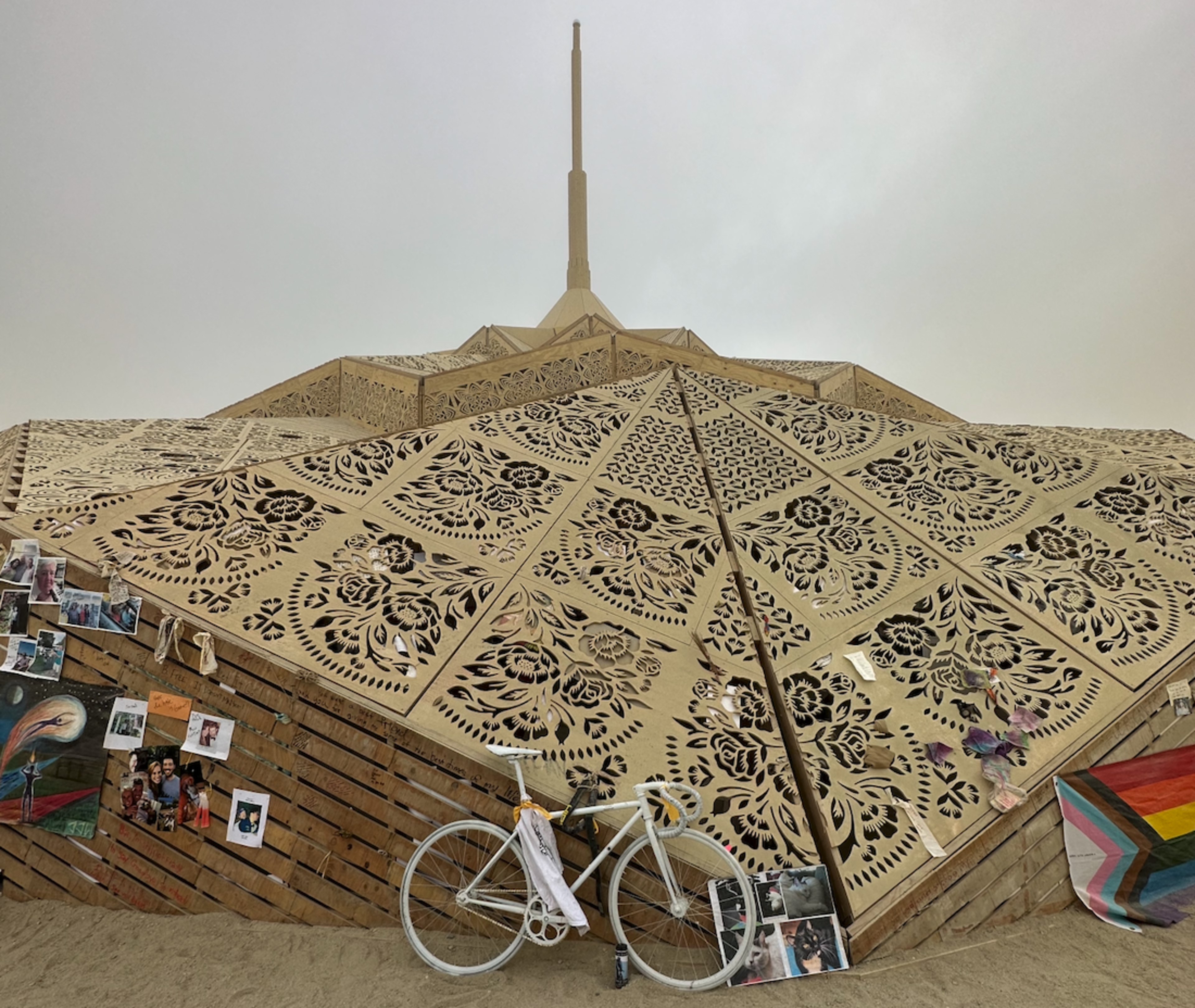 A large, intricately patterned wooden structure rises on sandy ground, decorated with photos, a white bicycle, and a colorful Pride flag on its side.