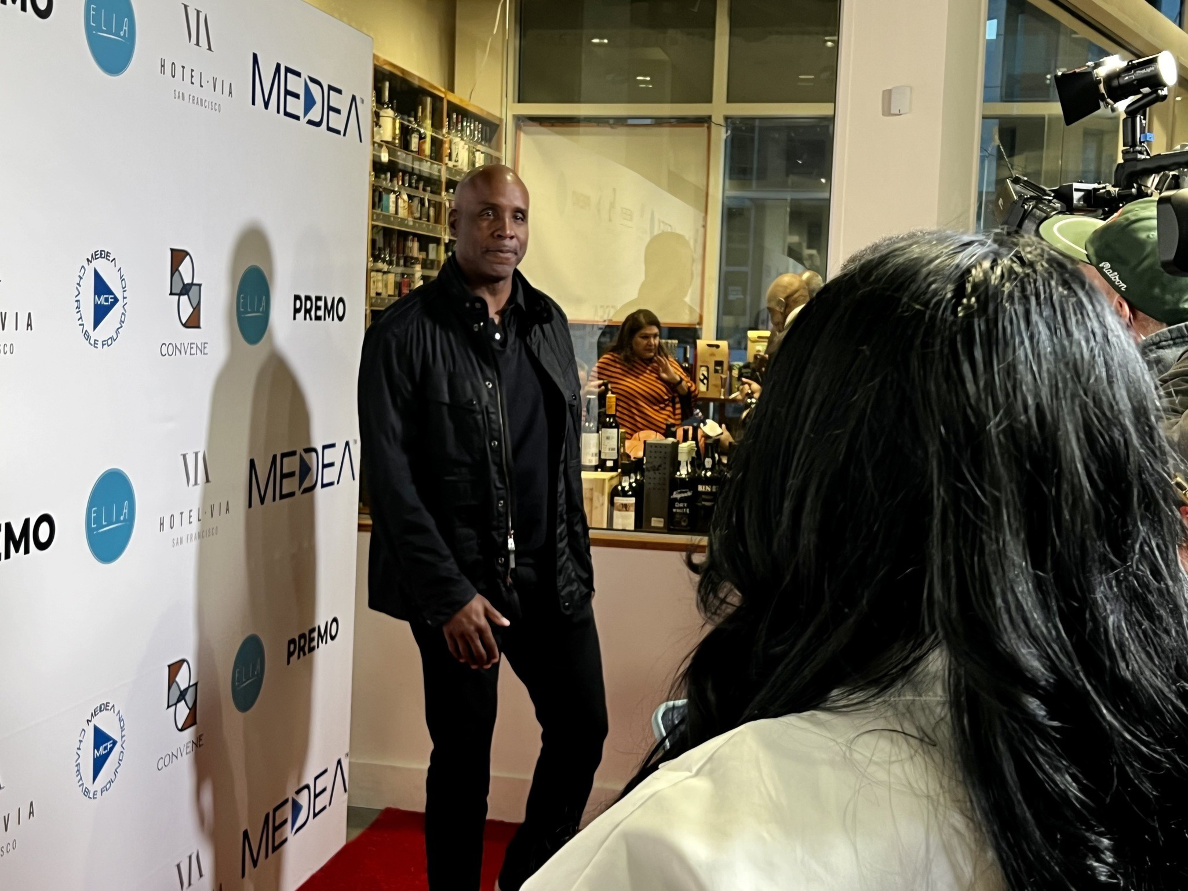 A man stands before a step-and-repeat banner at an event, with a camera crew and attendees nearby.