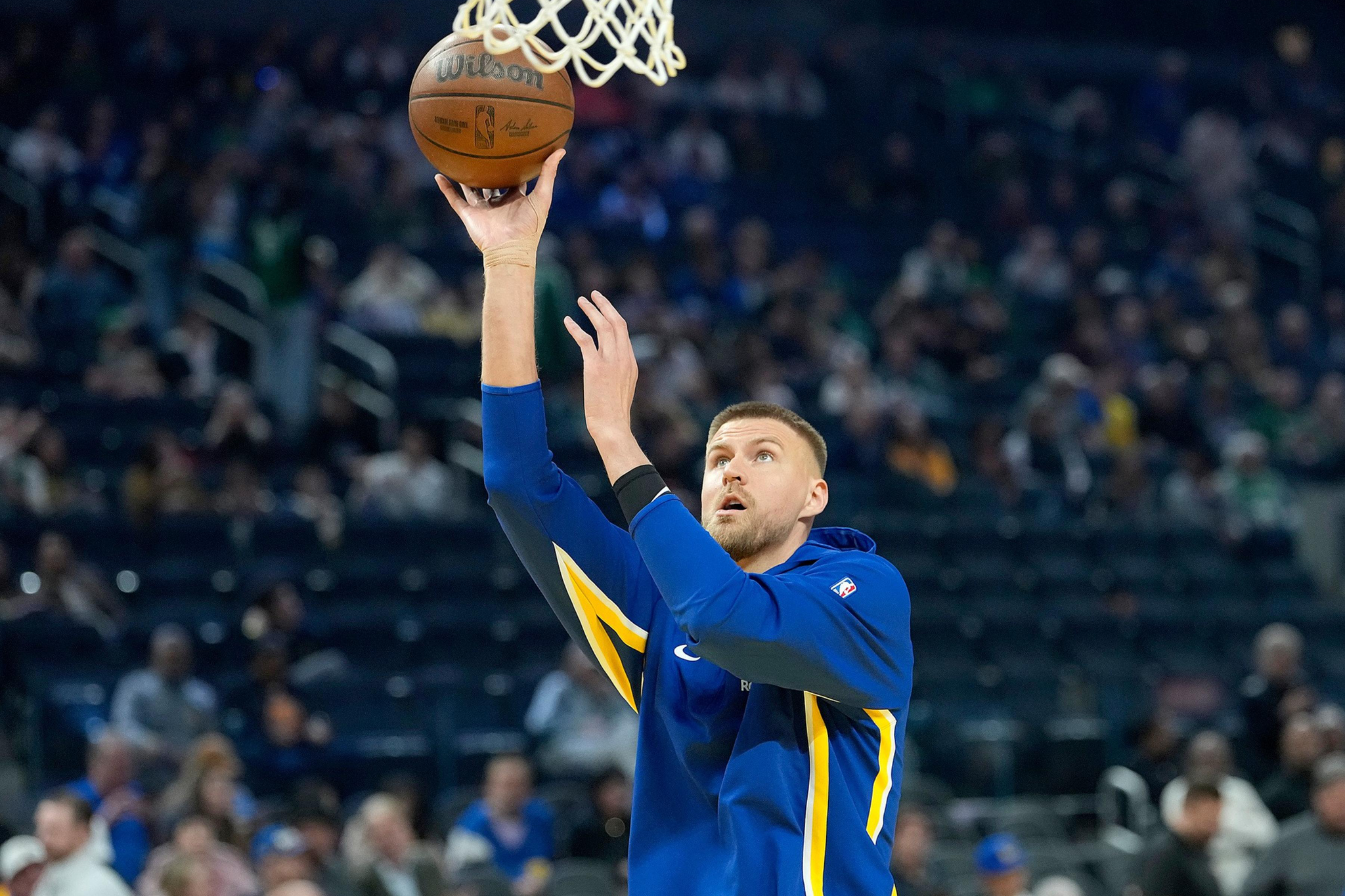 A basketball player in a blue and yellow warm-up jacket prepares to make a layup shot during a game with a crowd in the background.