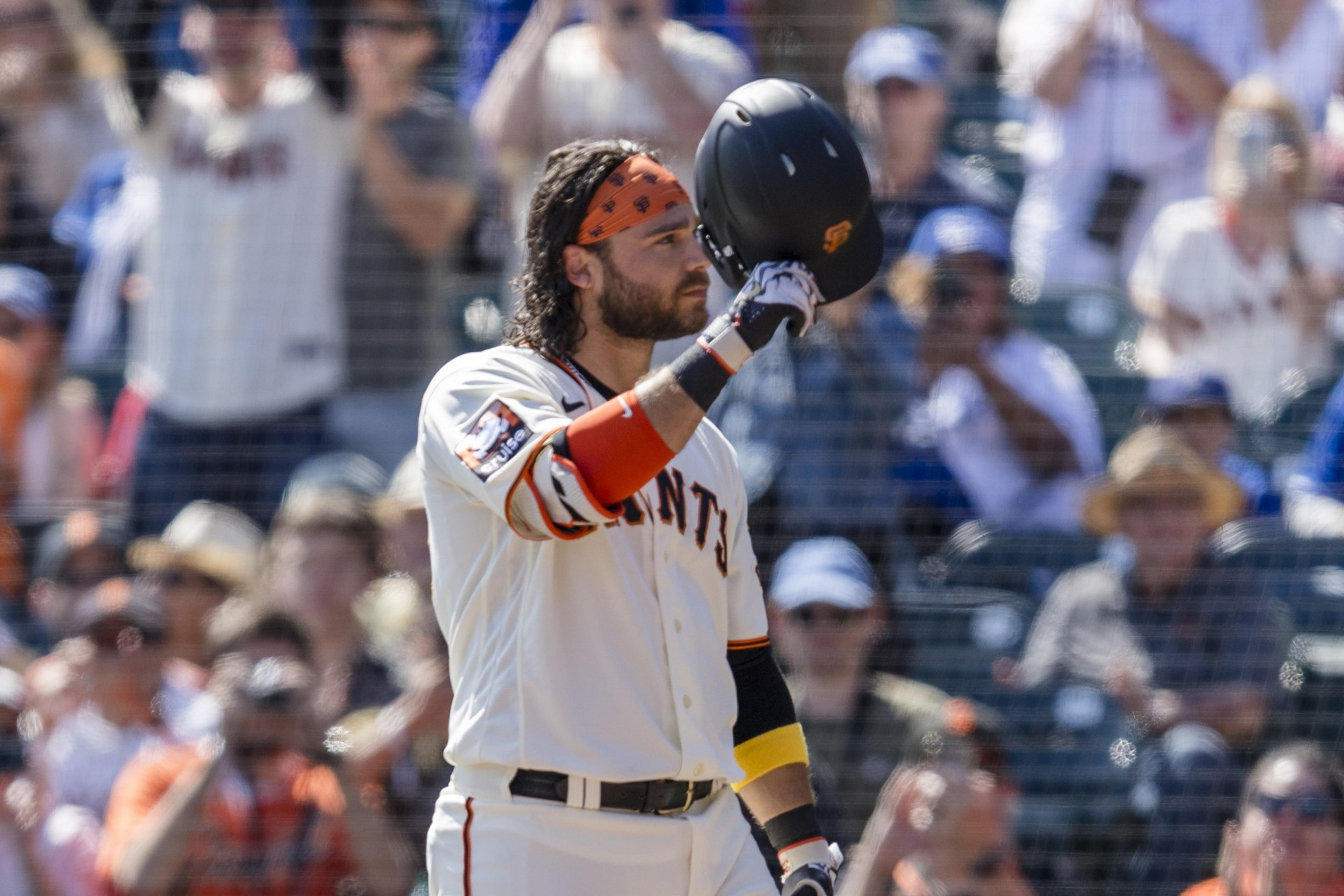 A baseball player in a white jersey with "Giants" holds his helmet to his lips. He wears a red bandana, wristbands, and the crowd is blurred in the background.