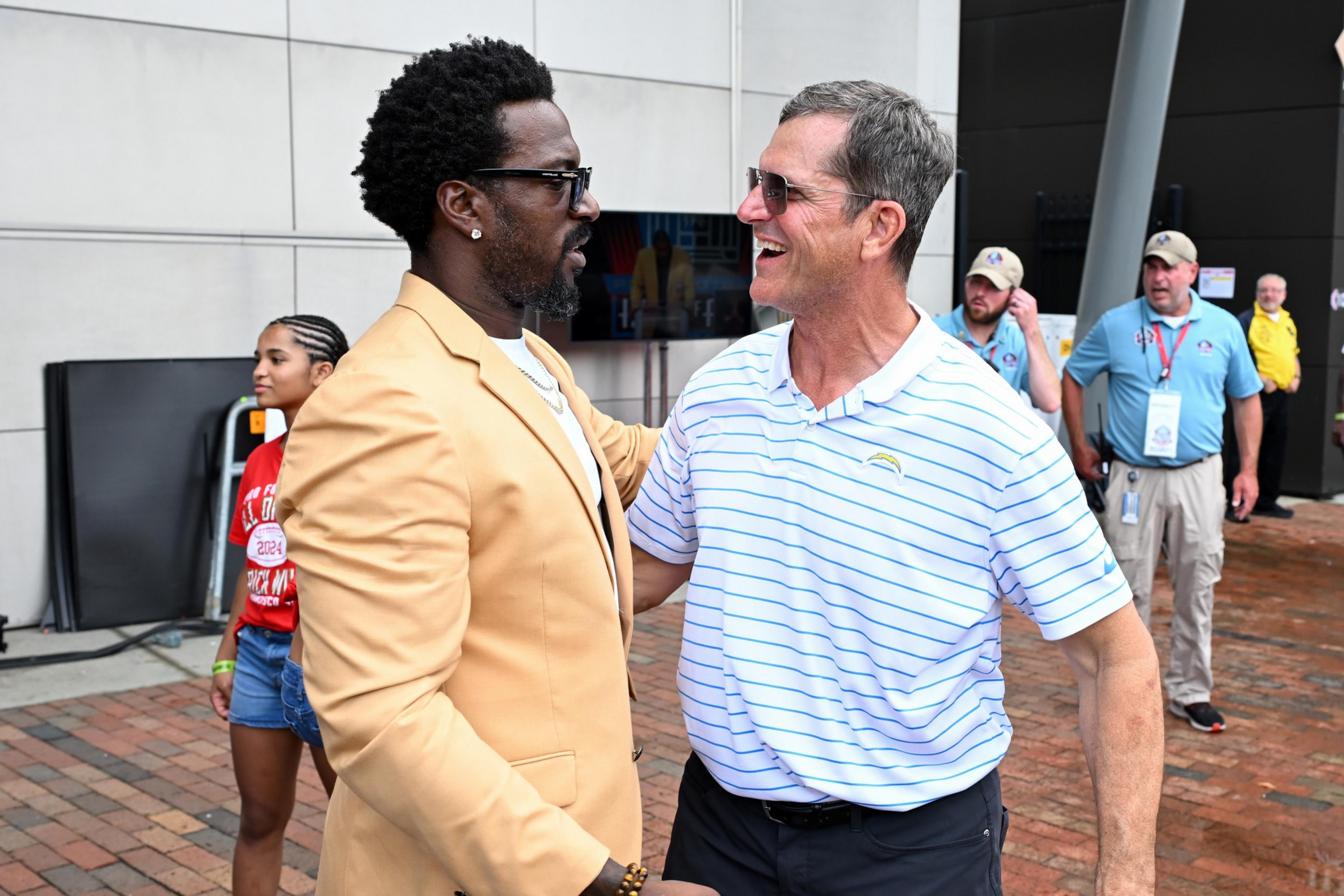 A man in a beige blazer and glasses embraces and smiles at a man in a white and blue striped polo shirt and sunglasses outside on a brick walkway.